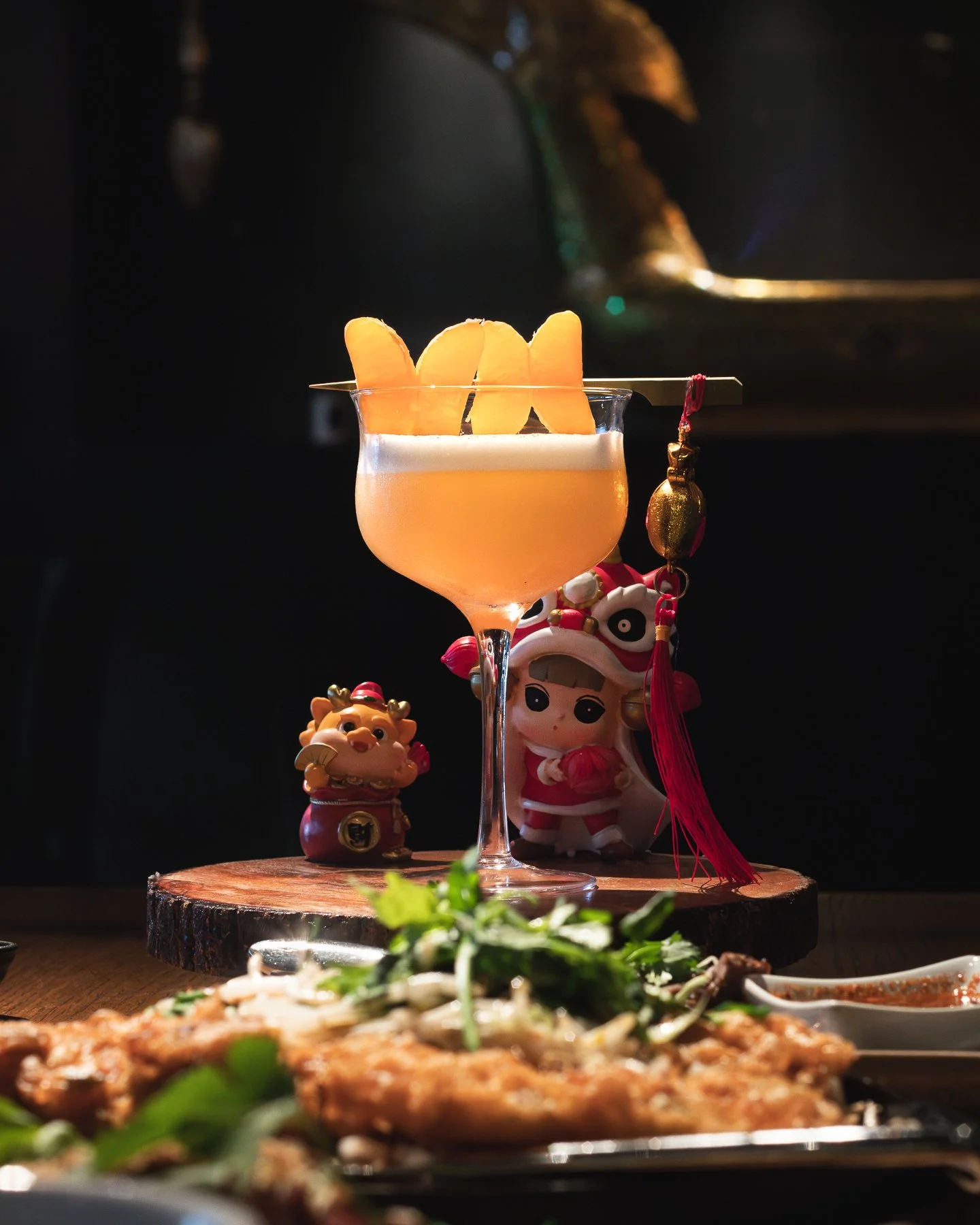 A cocktail from Bai Tong in Seattle garnished with orange slices on a skewer, placed on a wooden surface, with decorative figurines behind it and out-of-focus food in the foreground.