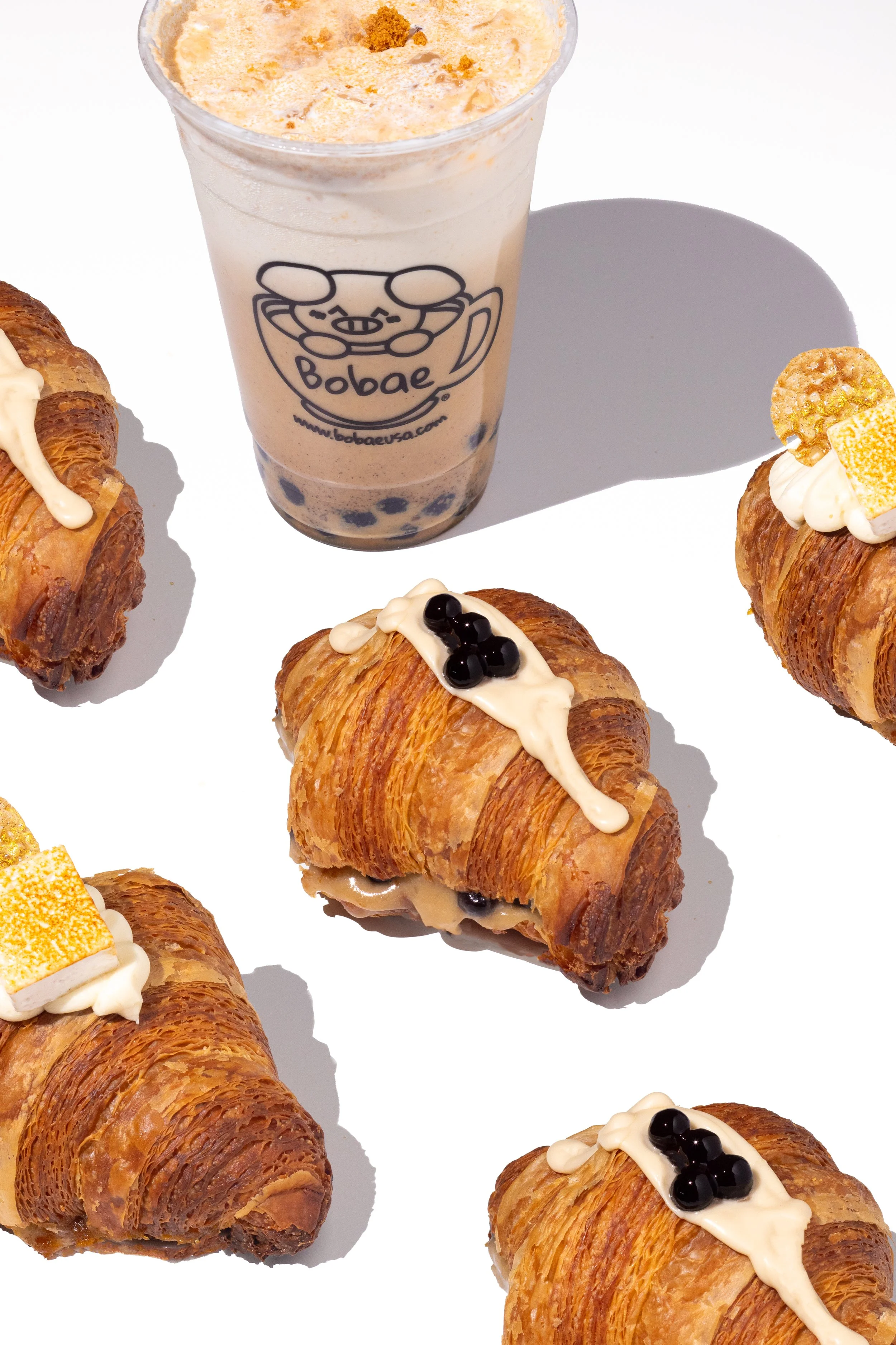 A flat lay of several croissants topped with different types of cream and toppings, surrounding a plastic cup of bubble tea with a pig cartoon logo from Bobae.