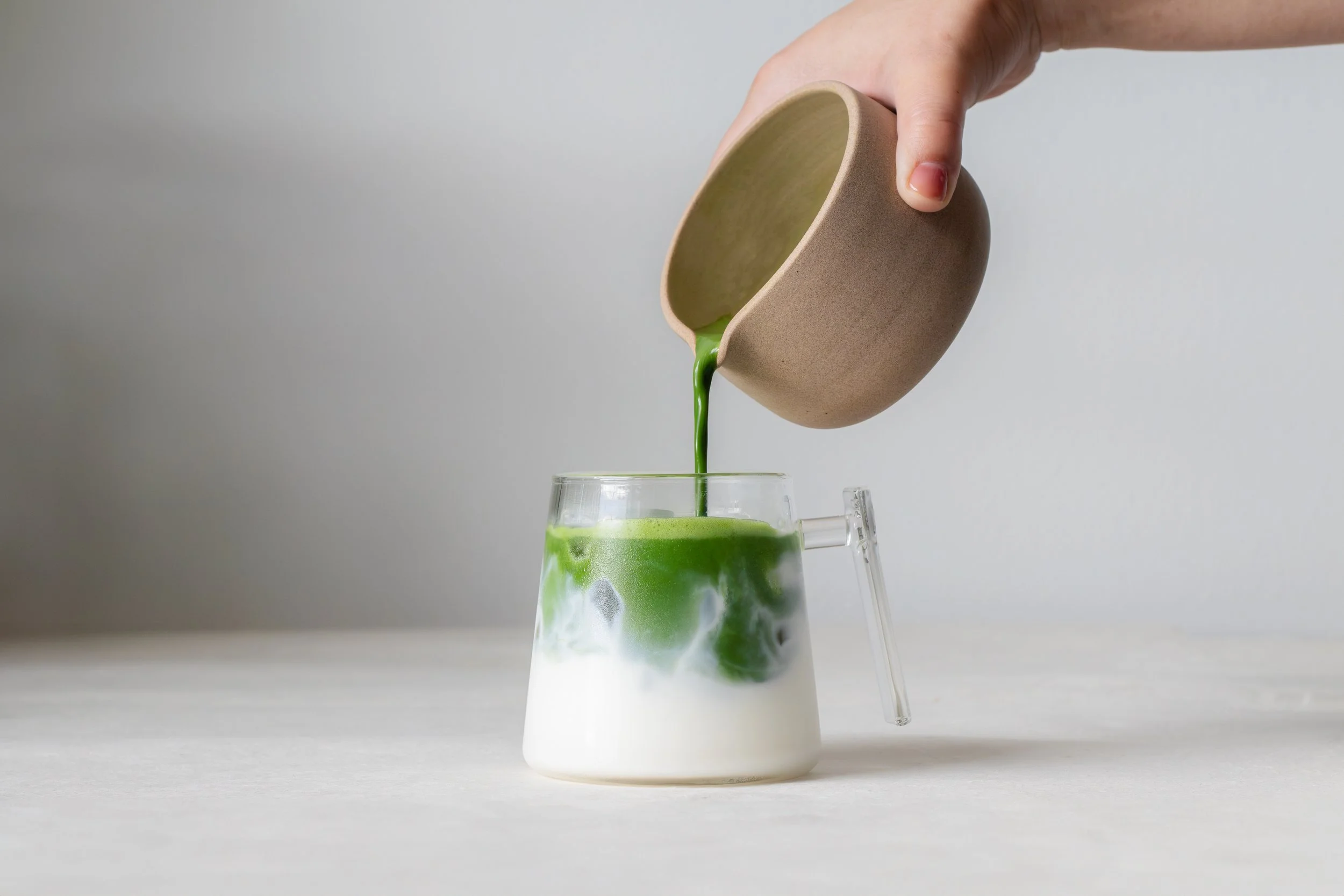 Hand pouring green matcha latte from a beige bowl into a glass with green and white swirling patterns.