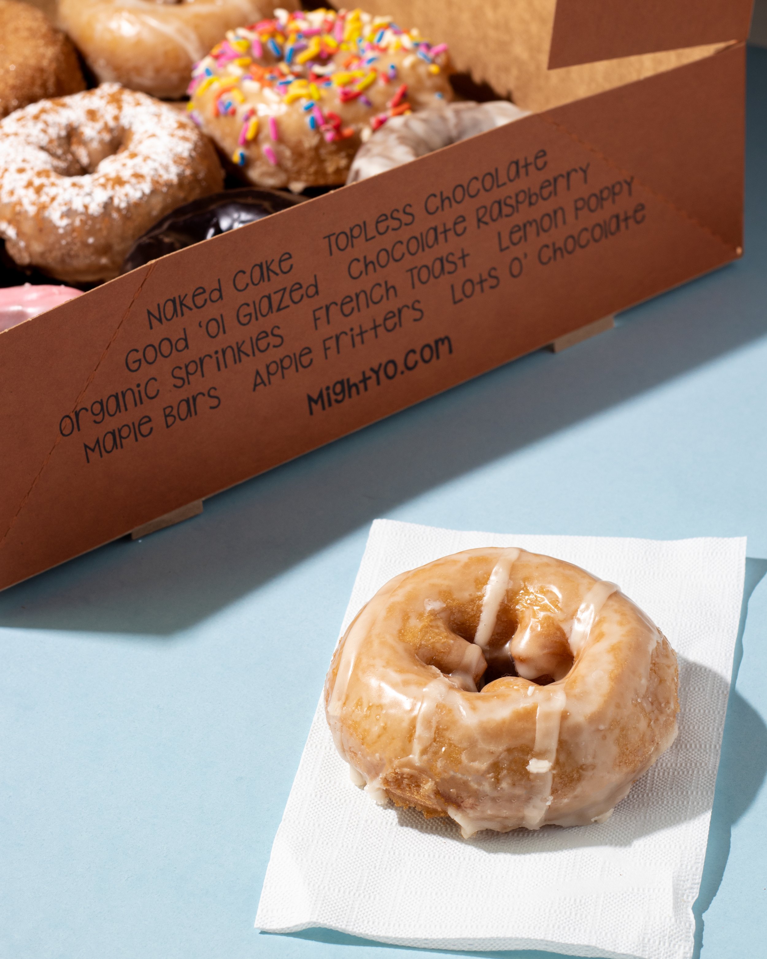 A glazed donut from Might-O with drizzle on a white napkin in the foreground, with a box of assorted donuts in the background, including sprinkles, powdered sugar, and chocolate glazed varieties.