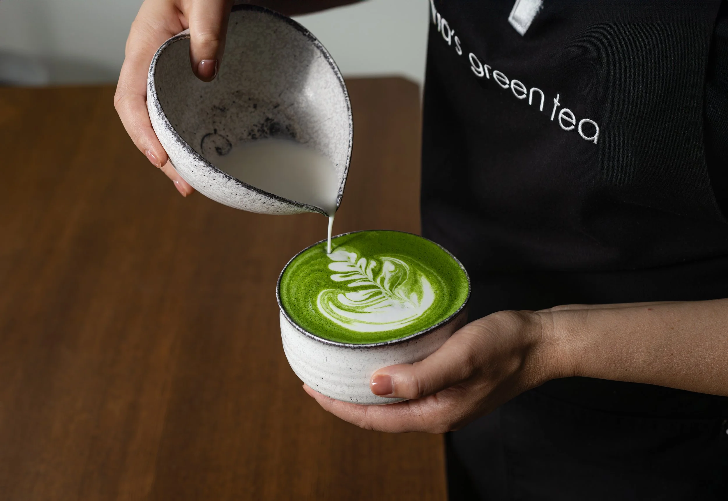A person pouring milk into a green matcha latte from Nana's Green Tea with latte art in a ceramic cup.