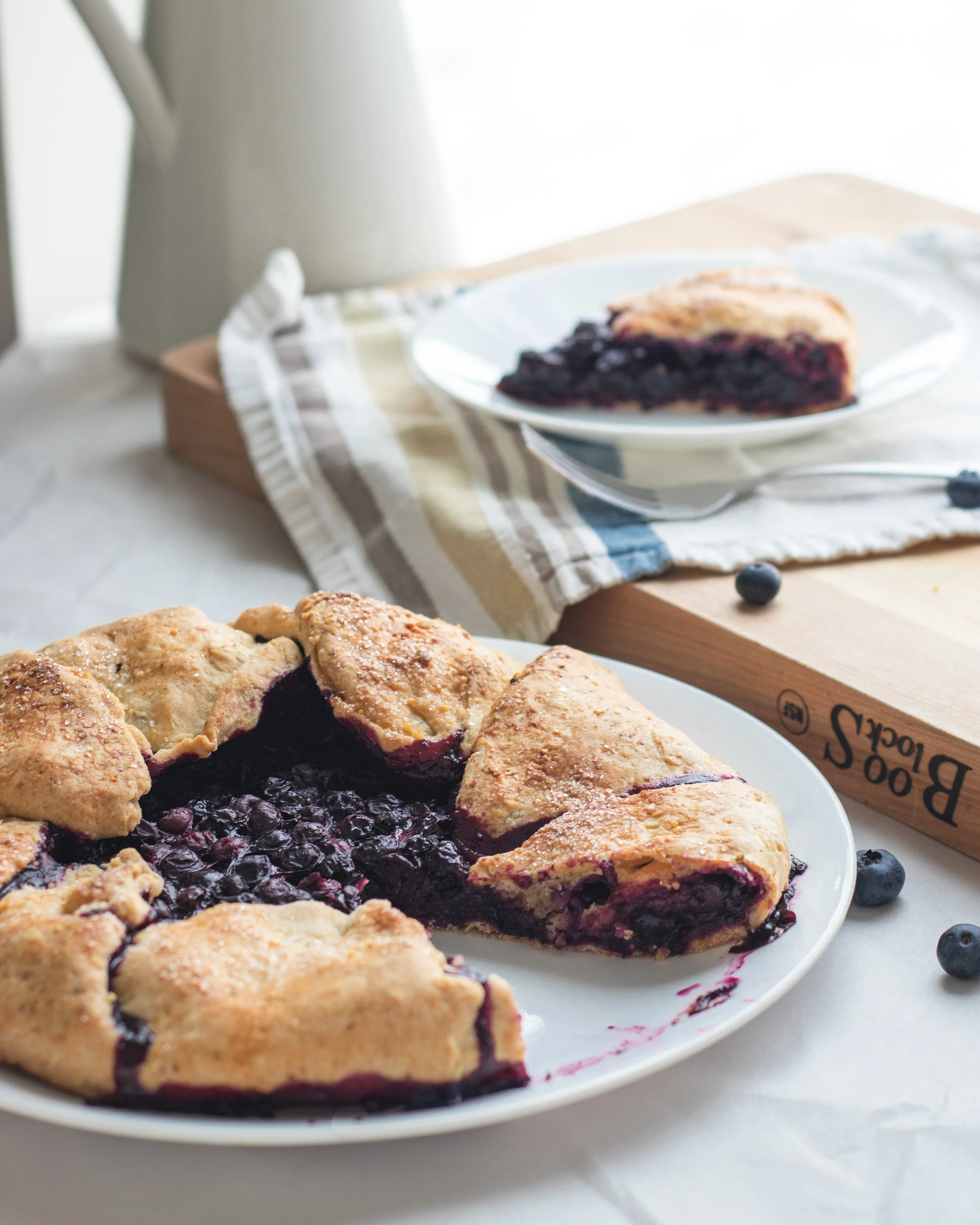 A blueberry pie with a golden baked crust on a white plate, with a fork beside it, placed on a white surface with natural lighting.