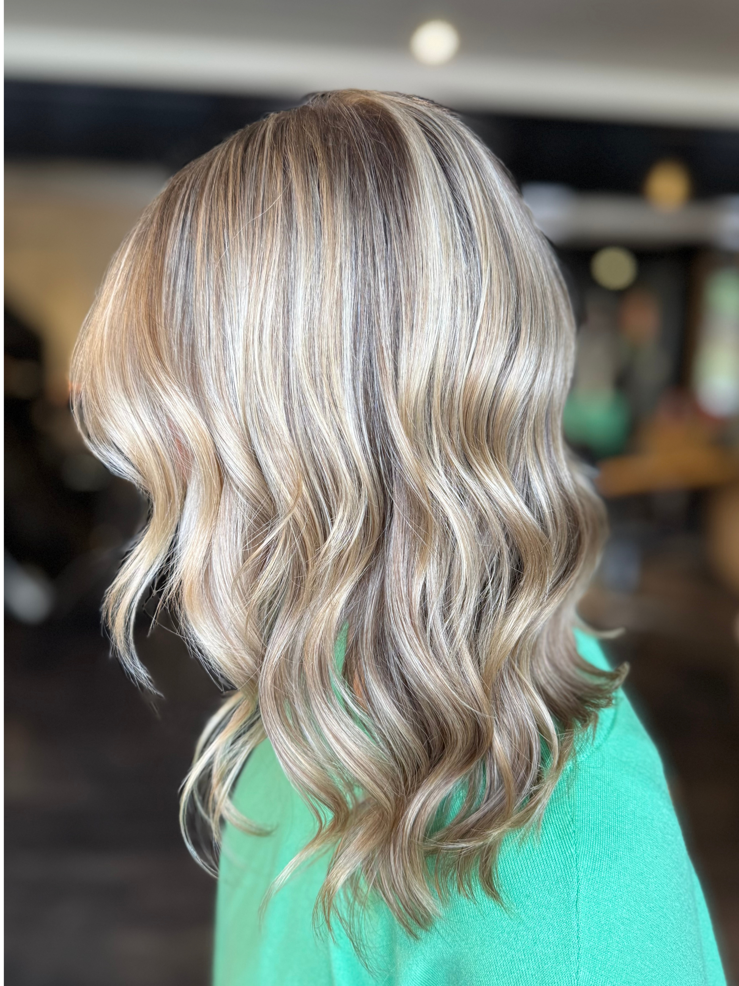 Back view of a woman with blonde wavy hair wearing a green shirt in an indoor setting.