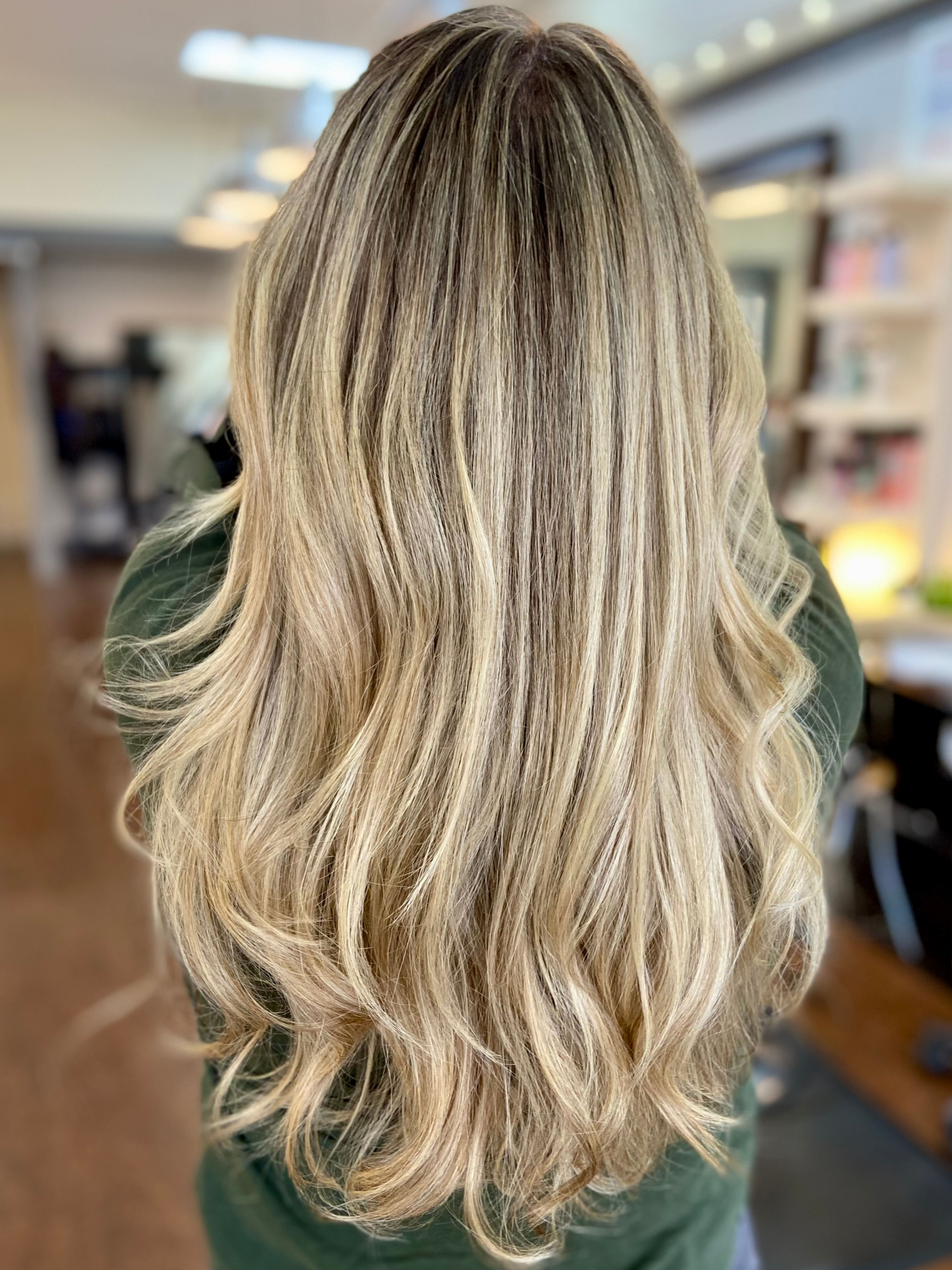 Back view of a person with long, wavy blonde hair inside a room with bookshelves and lighting