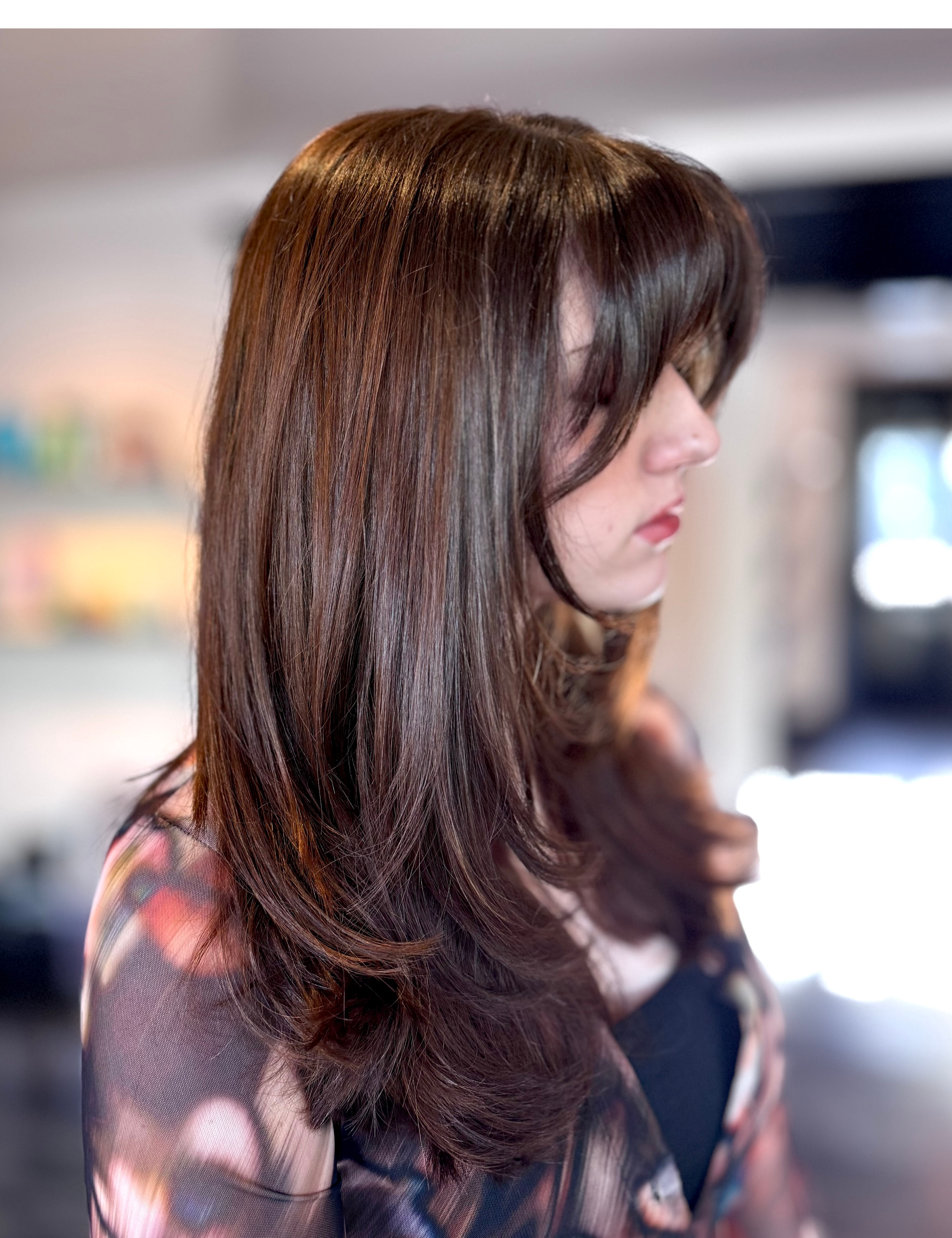 Side profile of a woman with long, wavy, dark brown hair, wearing a floral-patterned top, indoors with a blurred background.
