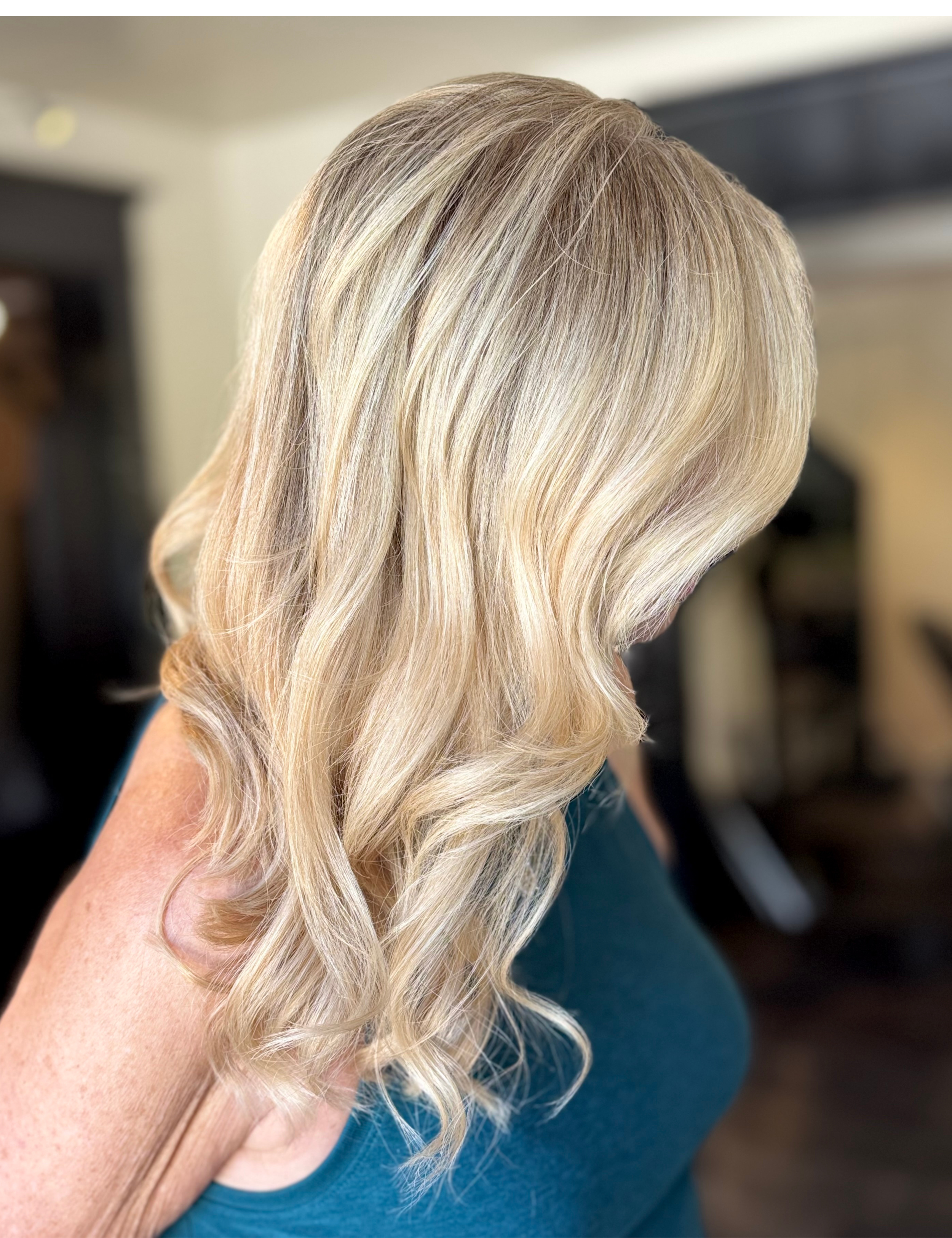 A close-up of a blonde woman's wavy hair with highlights, taken from the side, with a blurred background.