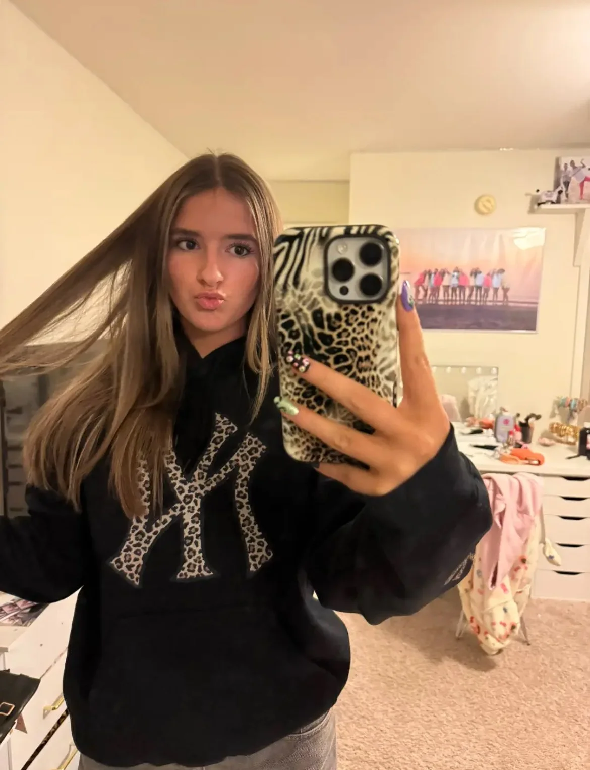 A young woman taking a mirror selfie with a smartphone in a bedroom, making a pouty face. She has long brown hair, light skin, and is wearing a black hoodie with a leopard print design on the front. The room in the background has a dresser with vario