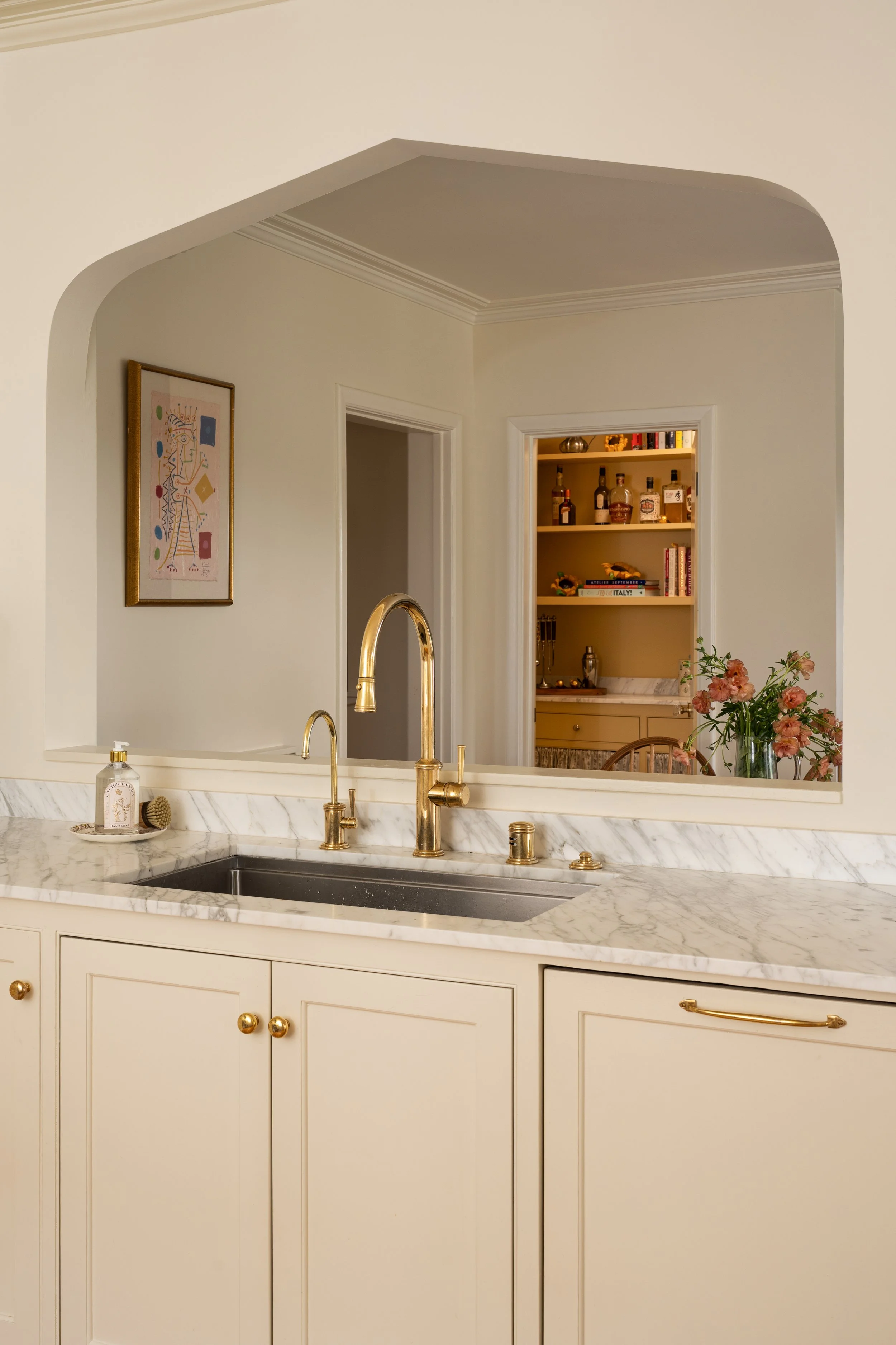 Moorish arch at brass faucet sink with marble counters looks into yellow cabinet pantry.