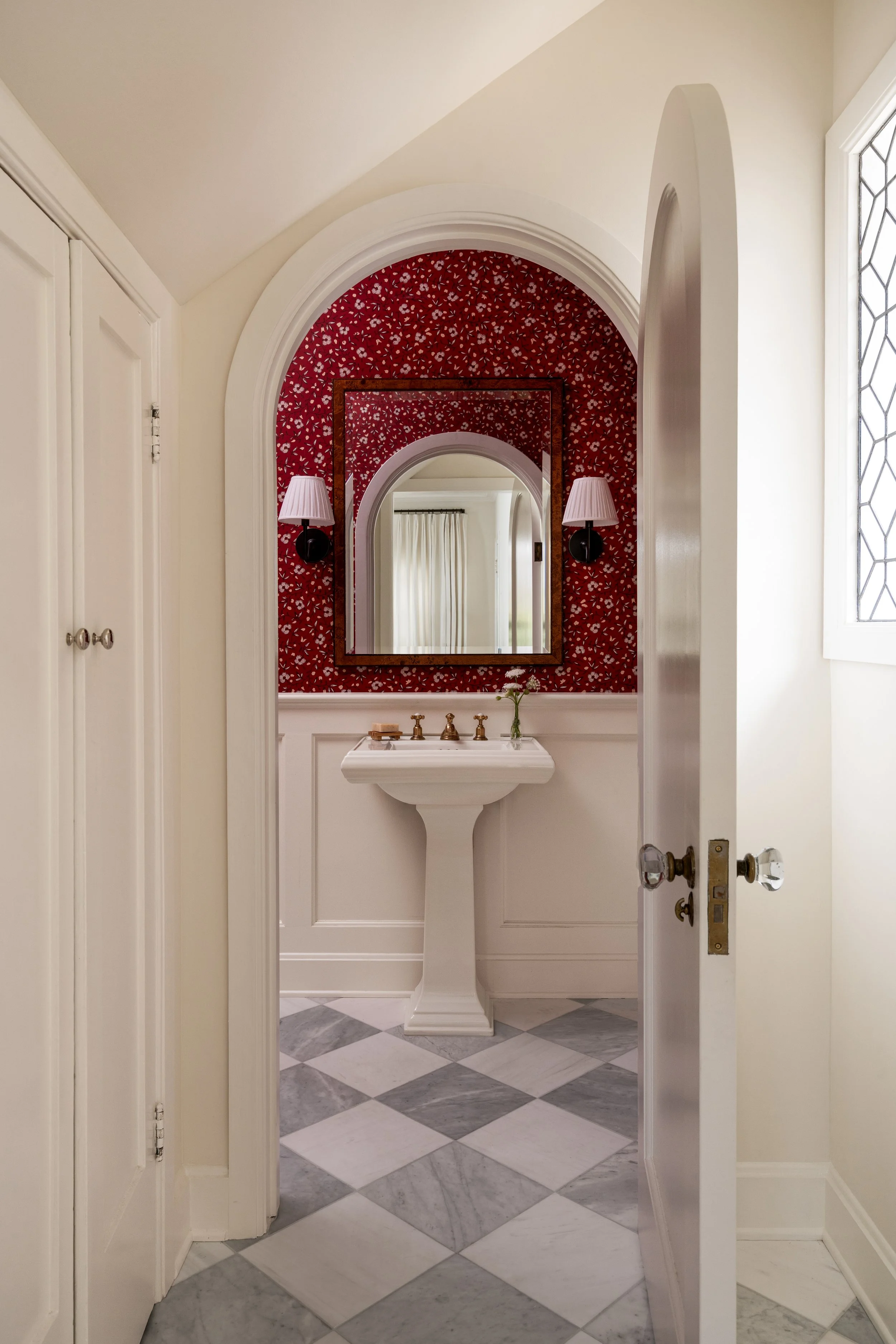 Arched door entry with leaded glass window to red wallpaper powder room and checkered marble floor.