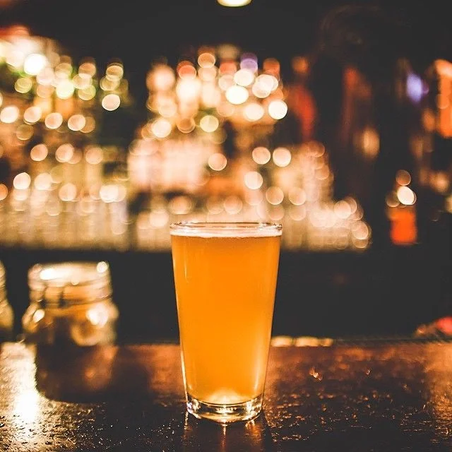 A glass of orange-colored beer on a bar counter with blurred colorful lights in the background.