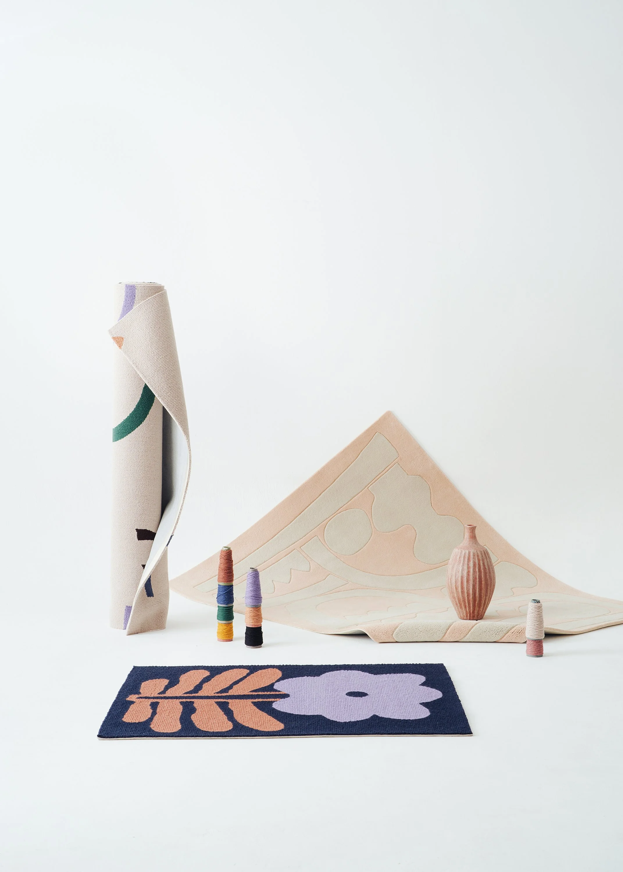 Arrangement of home decor items including a navy blue rug with a pink floral and leaf design, a beige rolled fabric, colorful thread spools, a pink striped vase, and abstract patterned fabric on a white background.