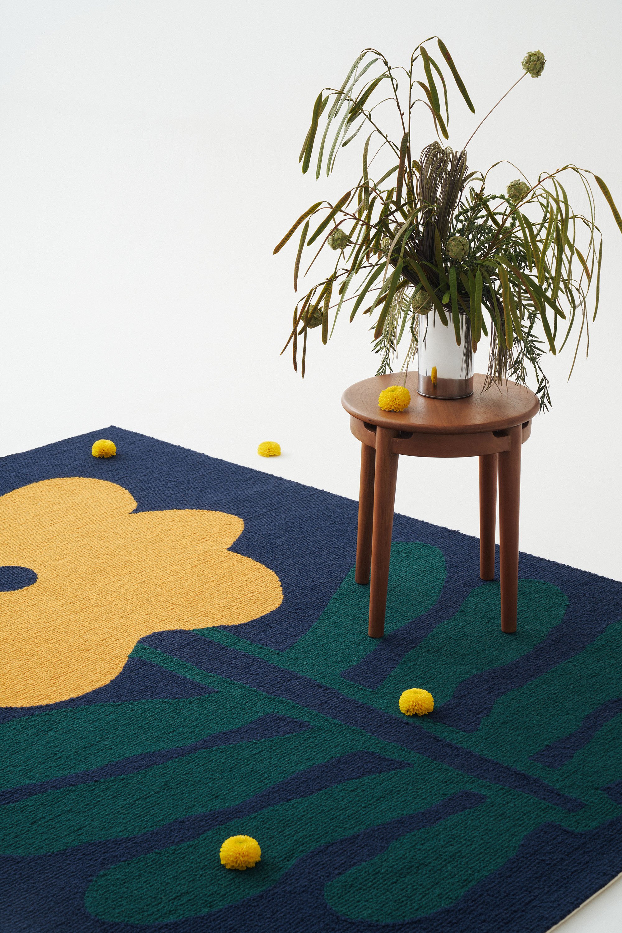 A plant with long green leaves and green spherical flowers in a glass vase on a small wooden table, with yellow flowers scattered on a colorful flower-shaped rug with green and dark blue sections against a white background.