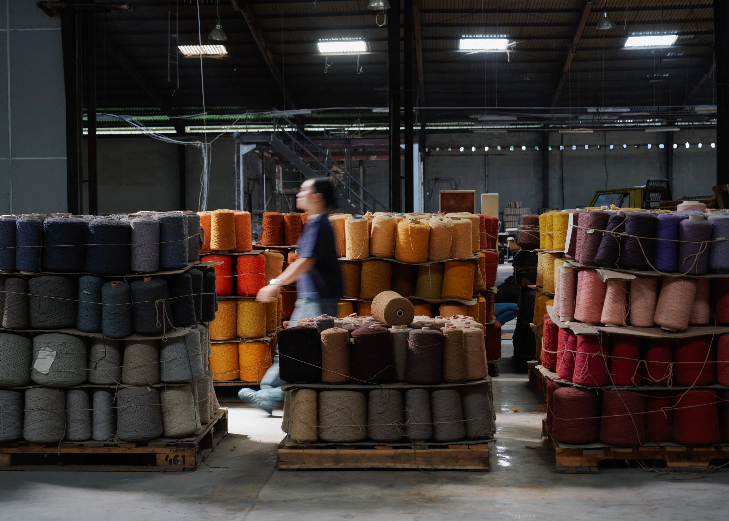 A person walking past stacks of colorful yarn spools in a factory or warehouse.