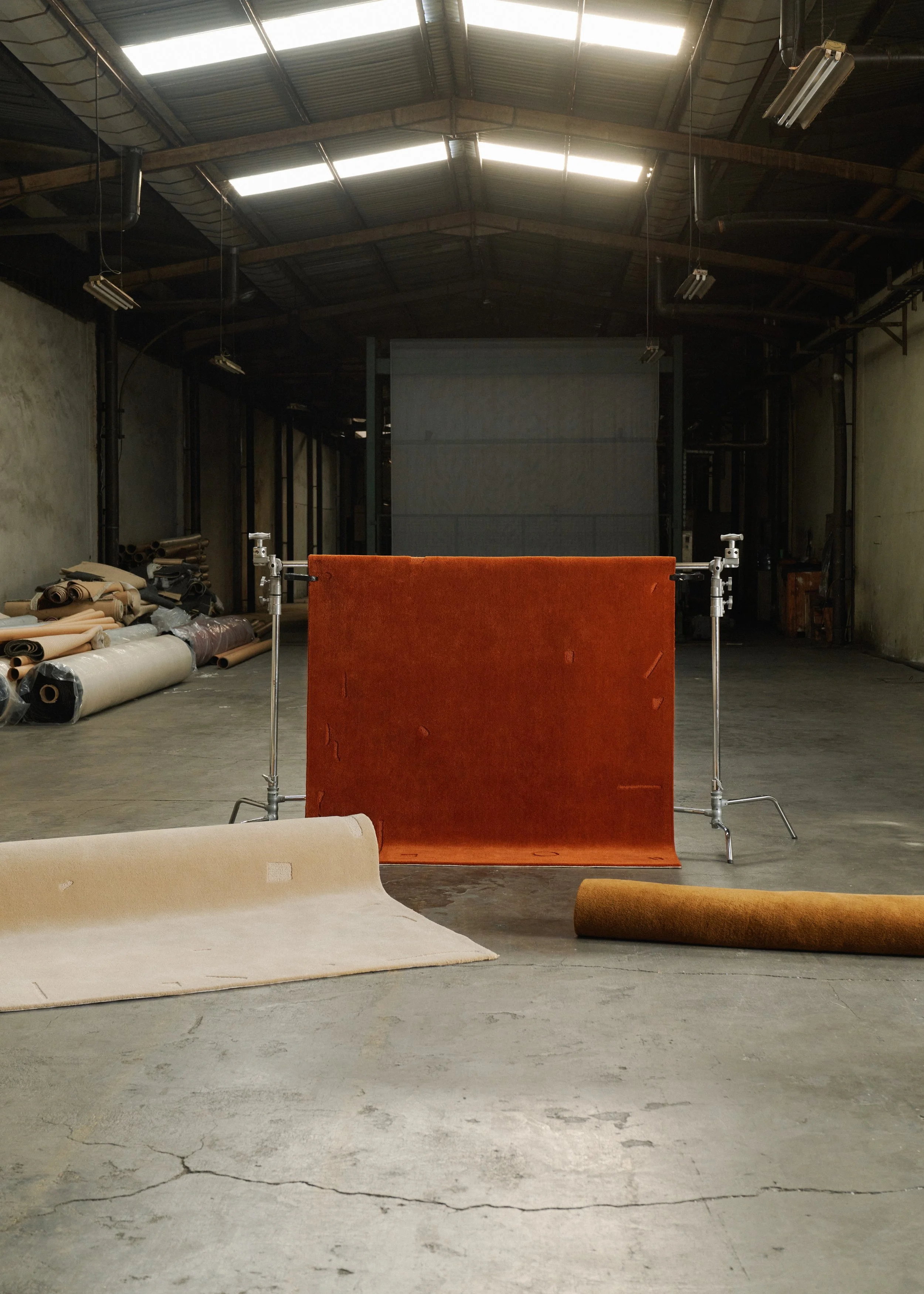 Empty industrial space with a large orange backdrop on metal stands, rolls of carpet and fabric on the floor, and a high, corrugated metal ceiling with sunlight coming through skylights.