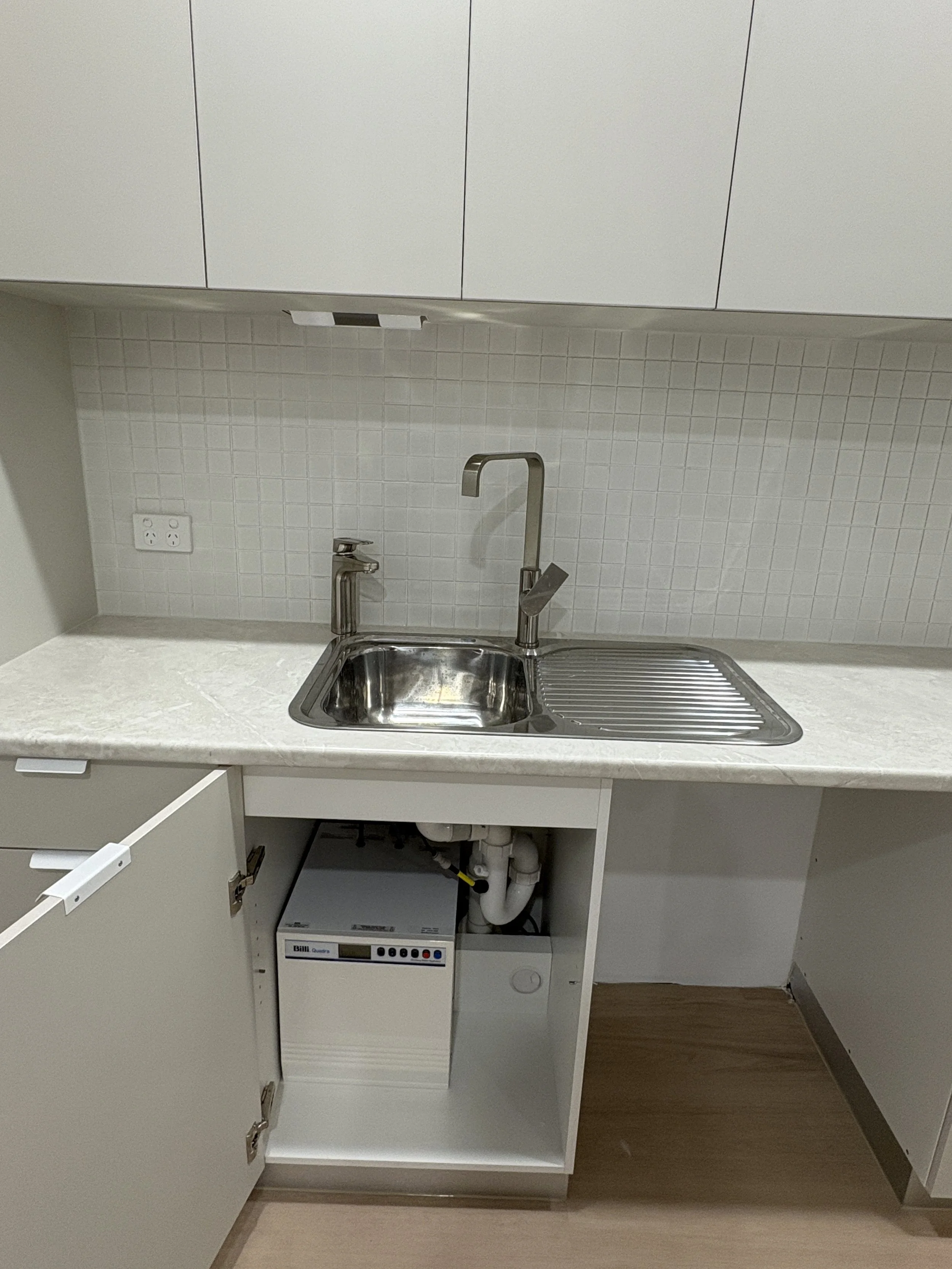 Kitchen with white cabinets, a stainless steel sink with two faucets, a white countertop, a tiled backsplash, and an open cabinet containing a white electronic device and pipes underneath the sink.
