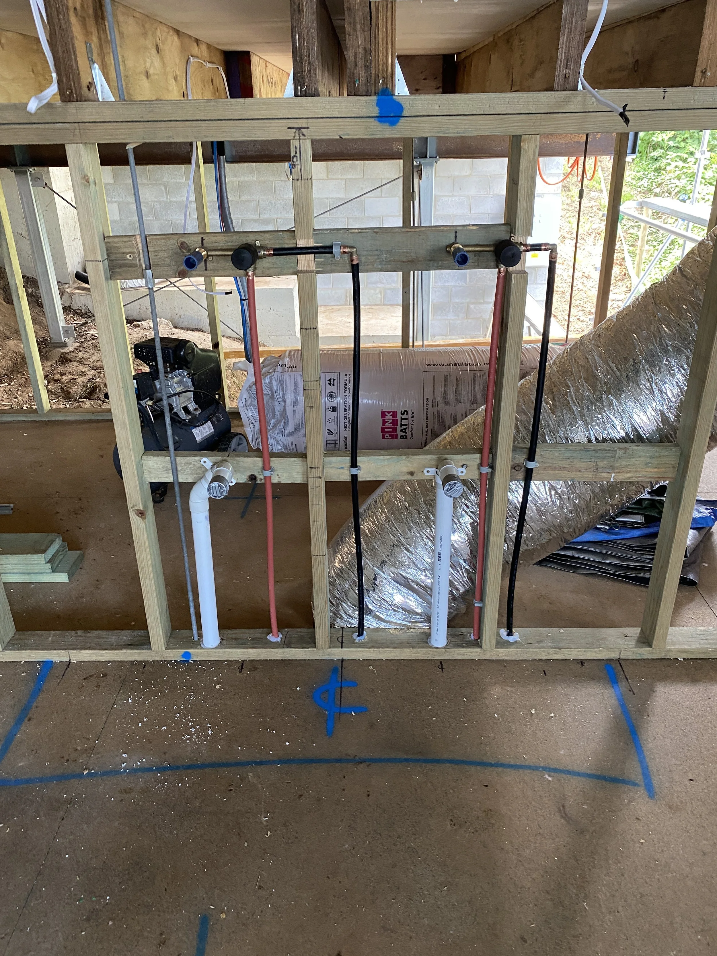 Construction site with exposed plumbing and framing for a bathroom or kitchen, showing pipe connections, an insulation roll, and a partially built wall.