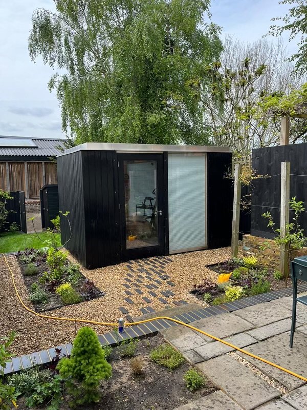 Small modern garden office with glass door in backyard, surrounded by gravel path and landscaped garden with plants and shrubs.