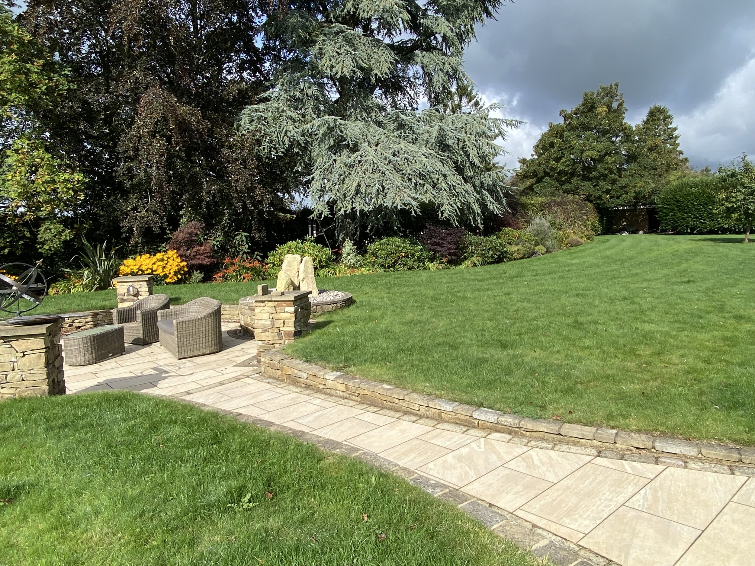 A landscaped garden with a stone patio, wicker chairs, and stone seating area surrounded by green grass and various trees and plants.