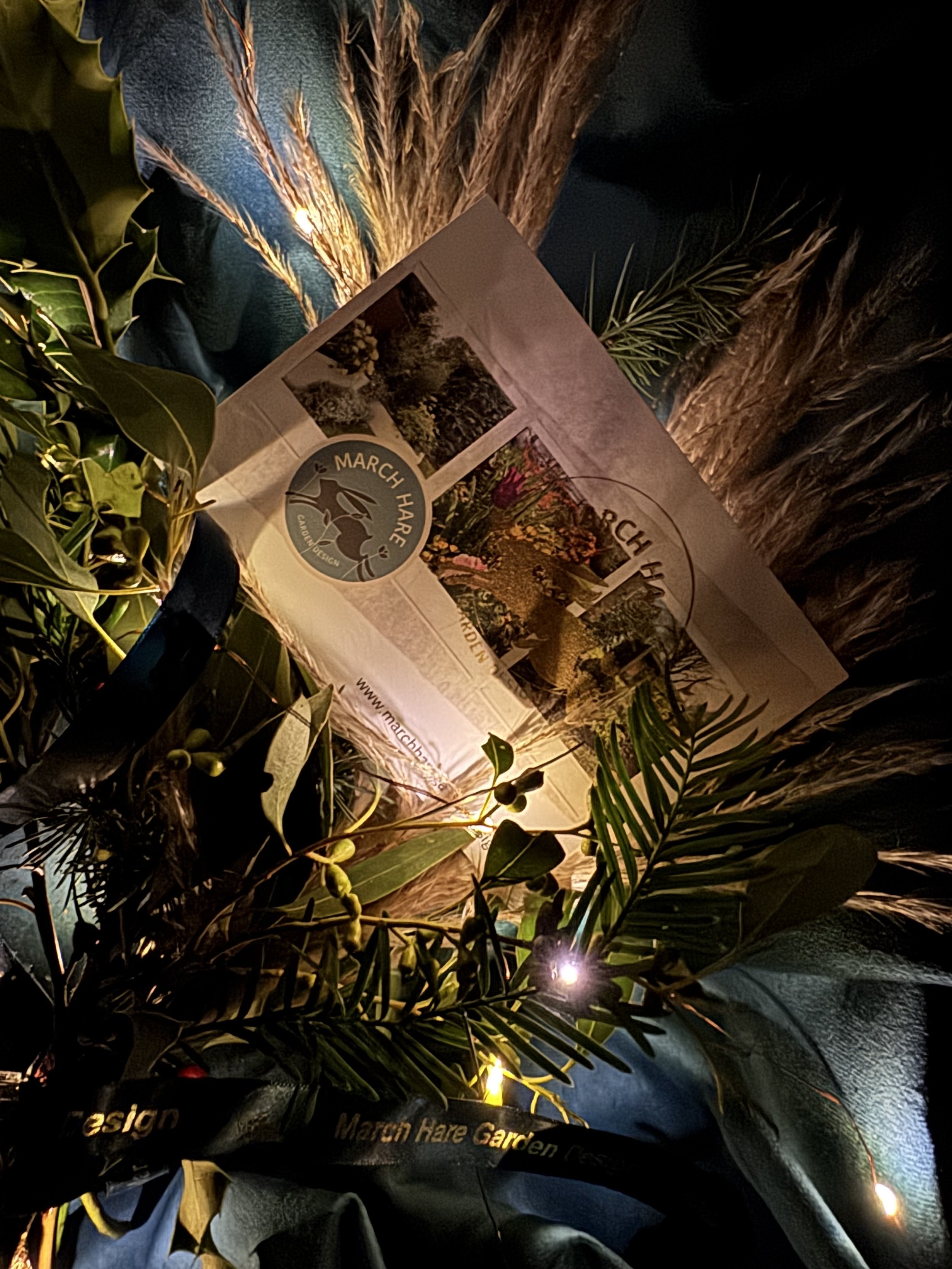 Assorted foliage and grasses surround a brochure for "March Hare Garden Design" against a teal fabric background, illuminated by small lights.
