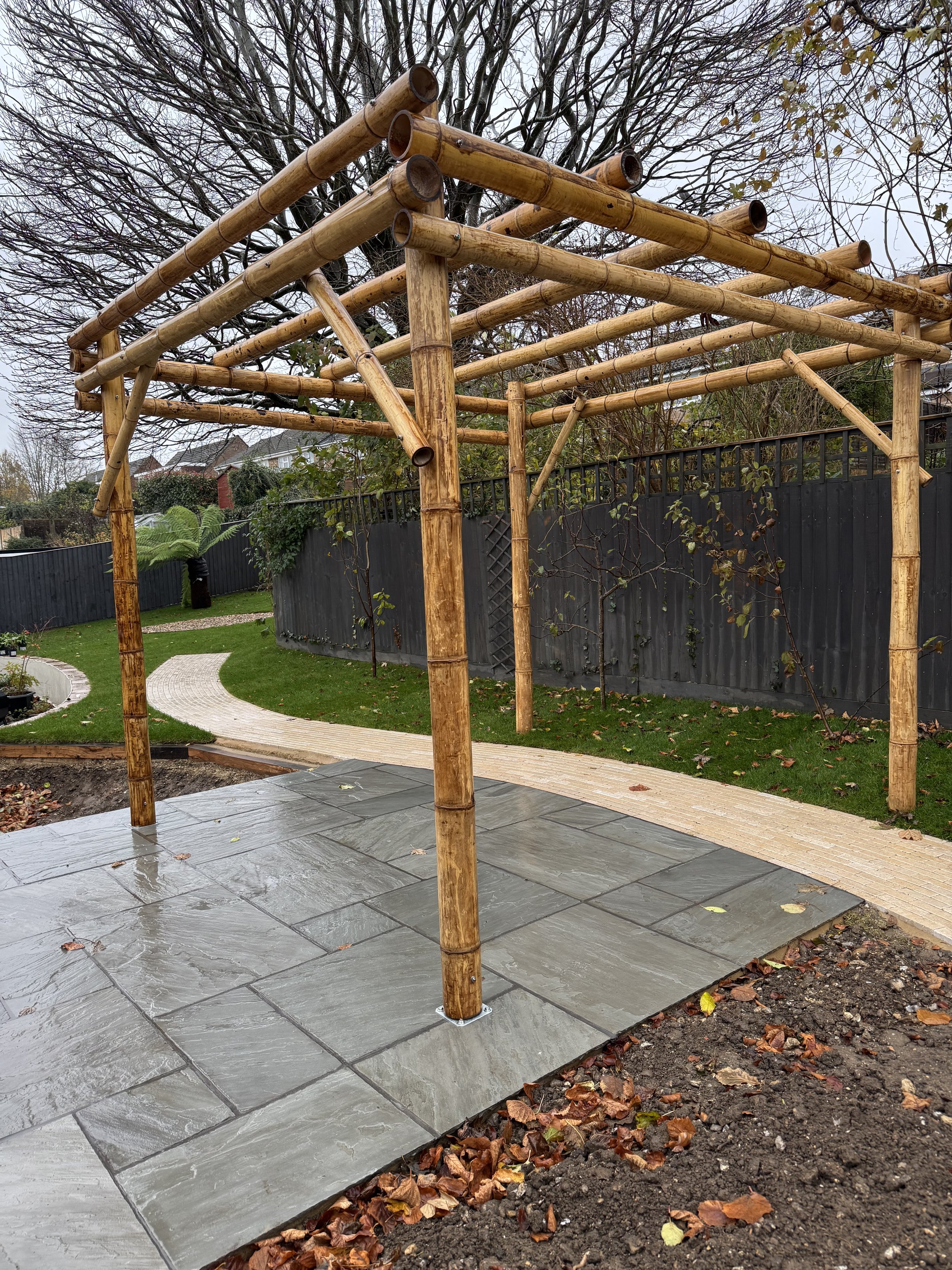 Wooden pergola with bamboo poles on a stone patio in a garden setting.