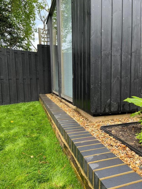Modern shed with dark siding next to grass and gravel, bordered by a low brick wall.