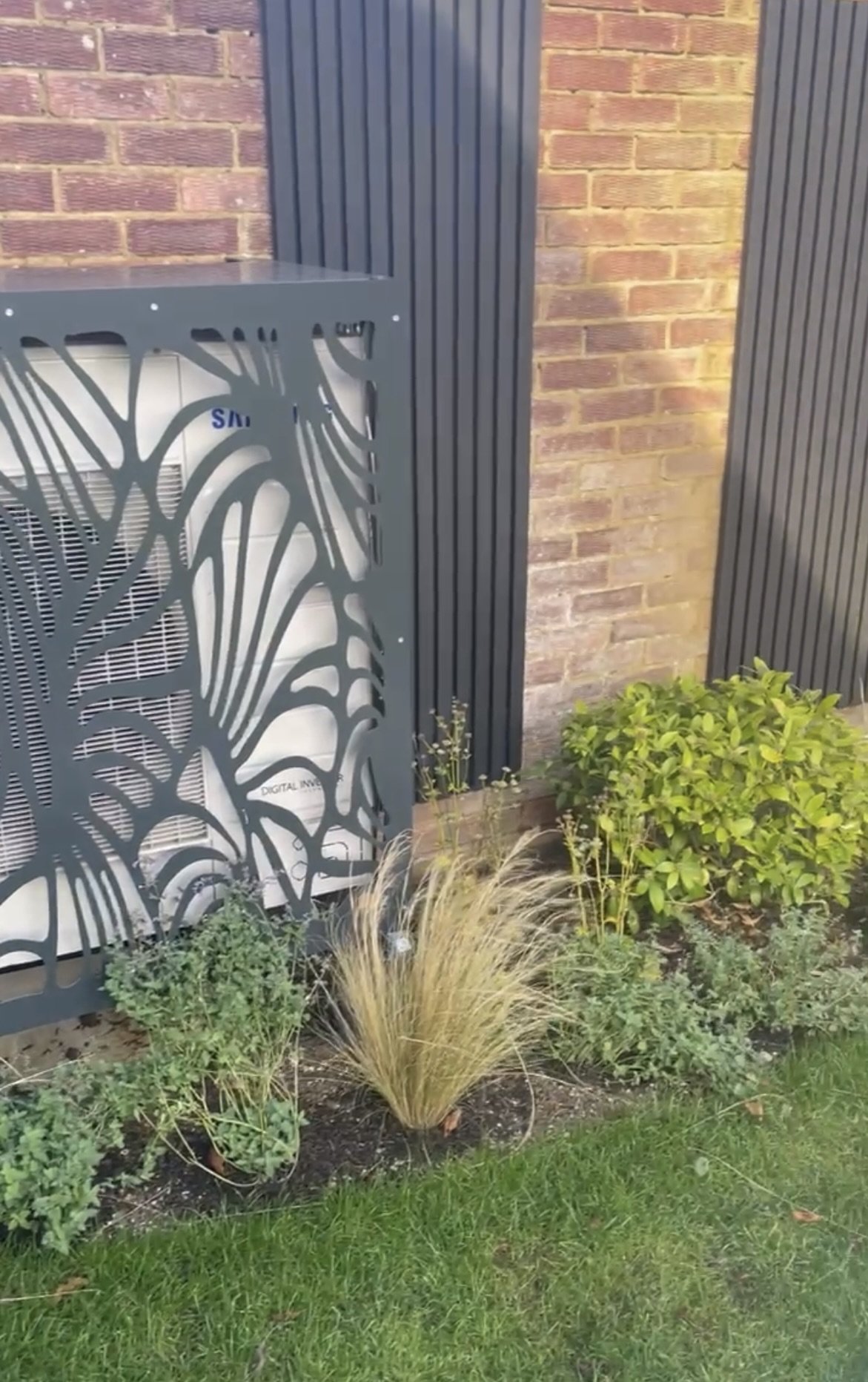 Decorative metal cover over outdoor air conditioning unit, surrounded by plants and grass, with brick wall and vertical panels in the background.