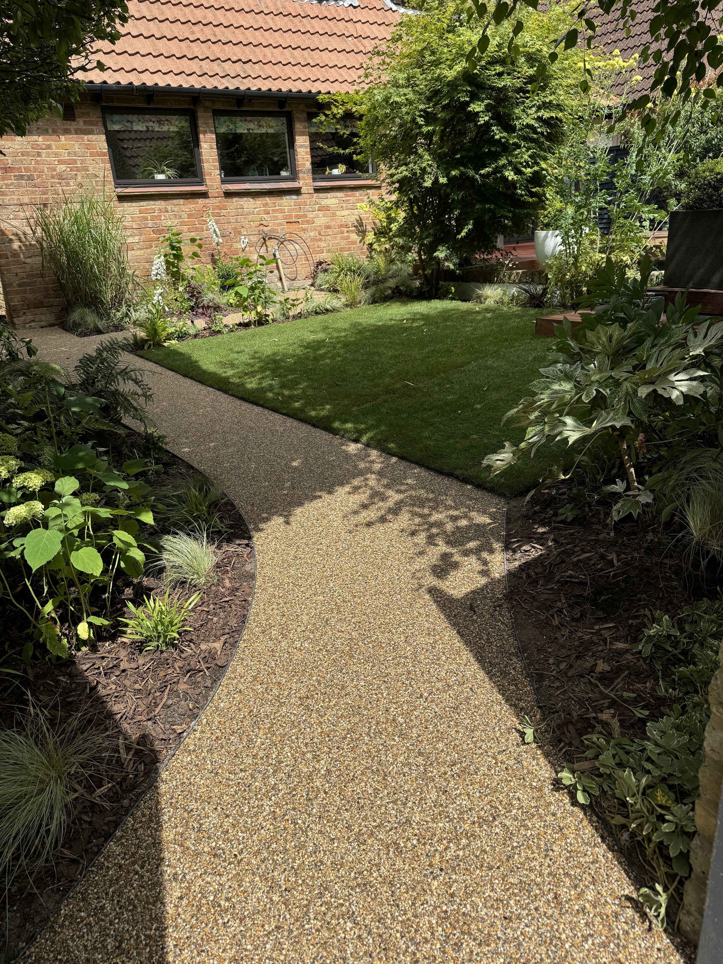 Garden with a curved gravel pathway leading to a brick house, surrounded by green plants and trees on both sides, under partly sunny skies.