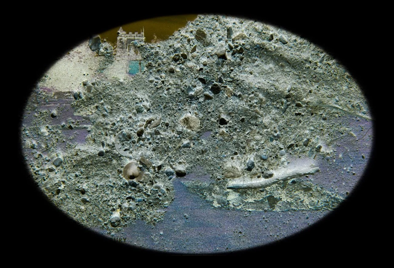 View through a telescope at a sandy, pebbled surface with scattered shells and small rocks, and a distant structure resembling a building or ruins in the background.
