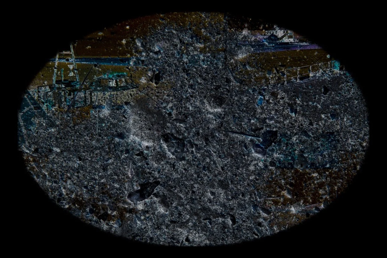 A view through a circular scope showing a rocky, uneven surface with some patches of debris and faint outlines of structures in the background.