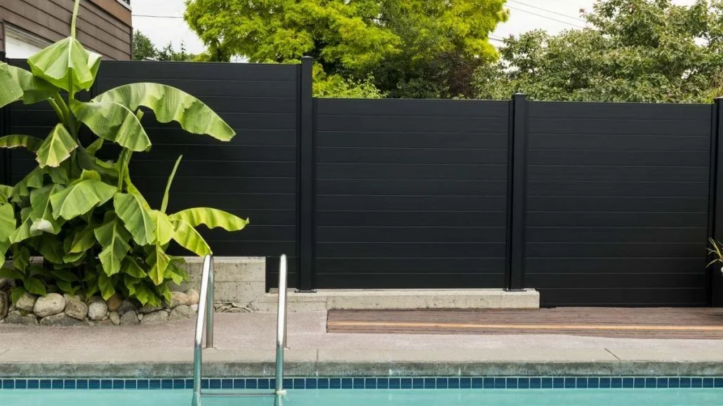 Outdoor pool with metal railing, adjacent to black horizontal slat fence, surrounded by green plants and trees.