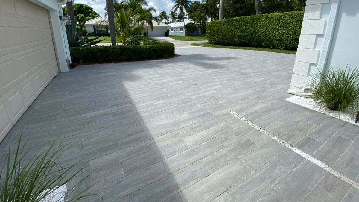 Modern driveway with light gray tiled surface, bordered by green shrubs, adjacent to a beige garage and white building facade.