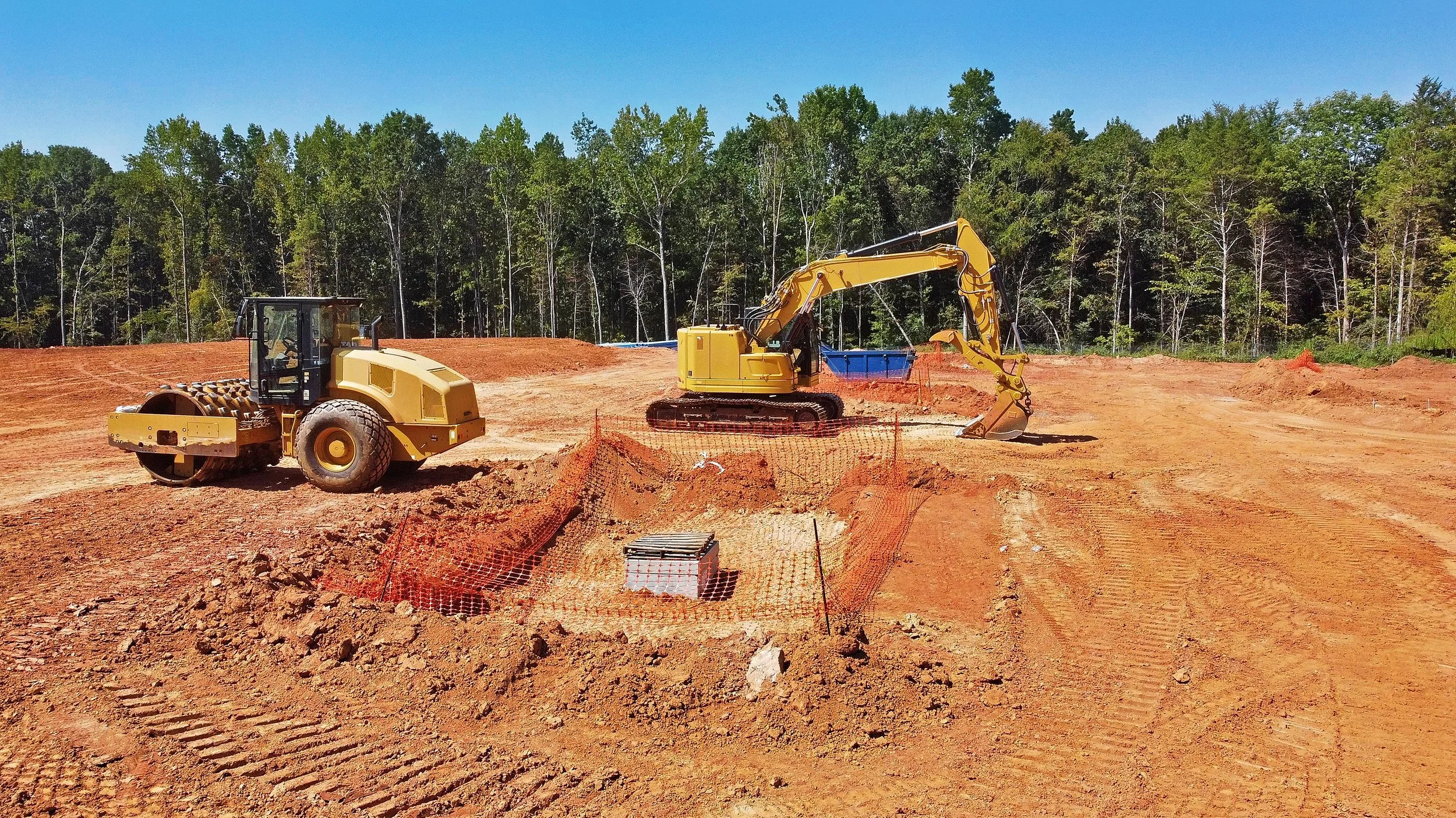 Construction site with bulldozer and backhoe on a dirt field surrounded by forest.
