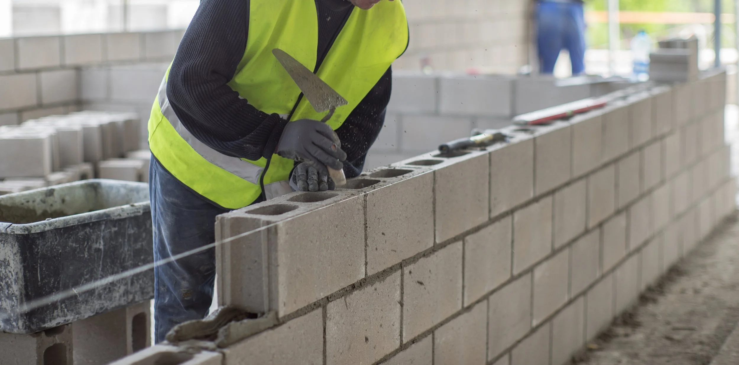 Construction worker laying cinder blocks using a trowel and mortar, wearing a yellow safety vest.