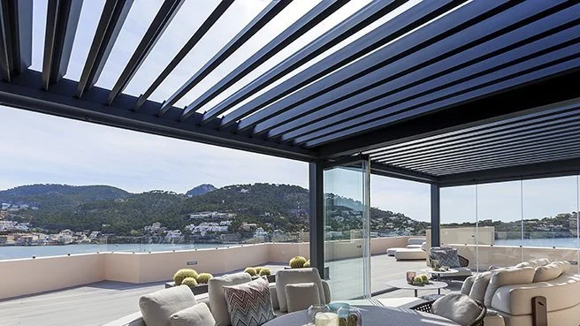 Modern outdoor terrace with sofas under a pergola, overlooking hills and waterfront.