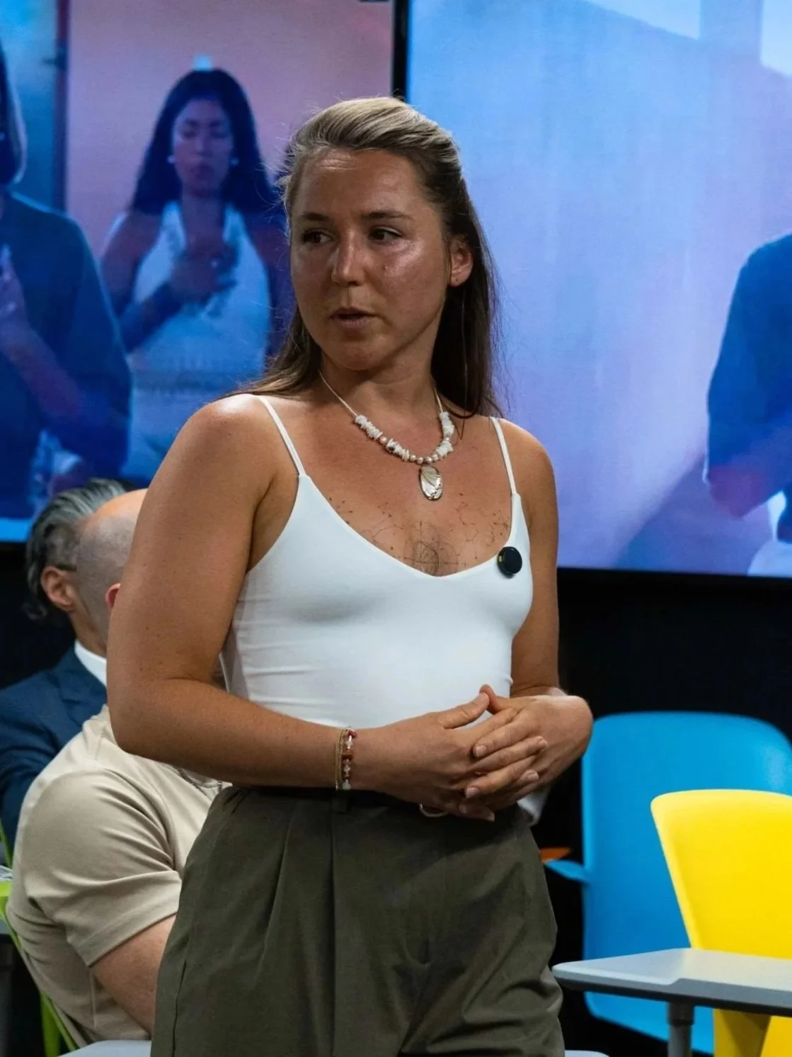 A woman standing in a room with colorful chairs, wearing a white spaghetti strap top and dark pants, speaking or presenting.