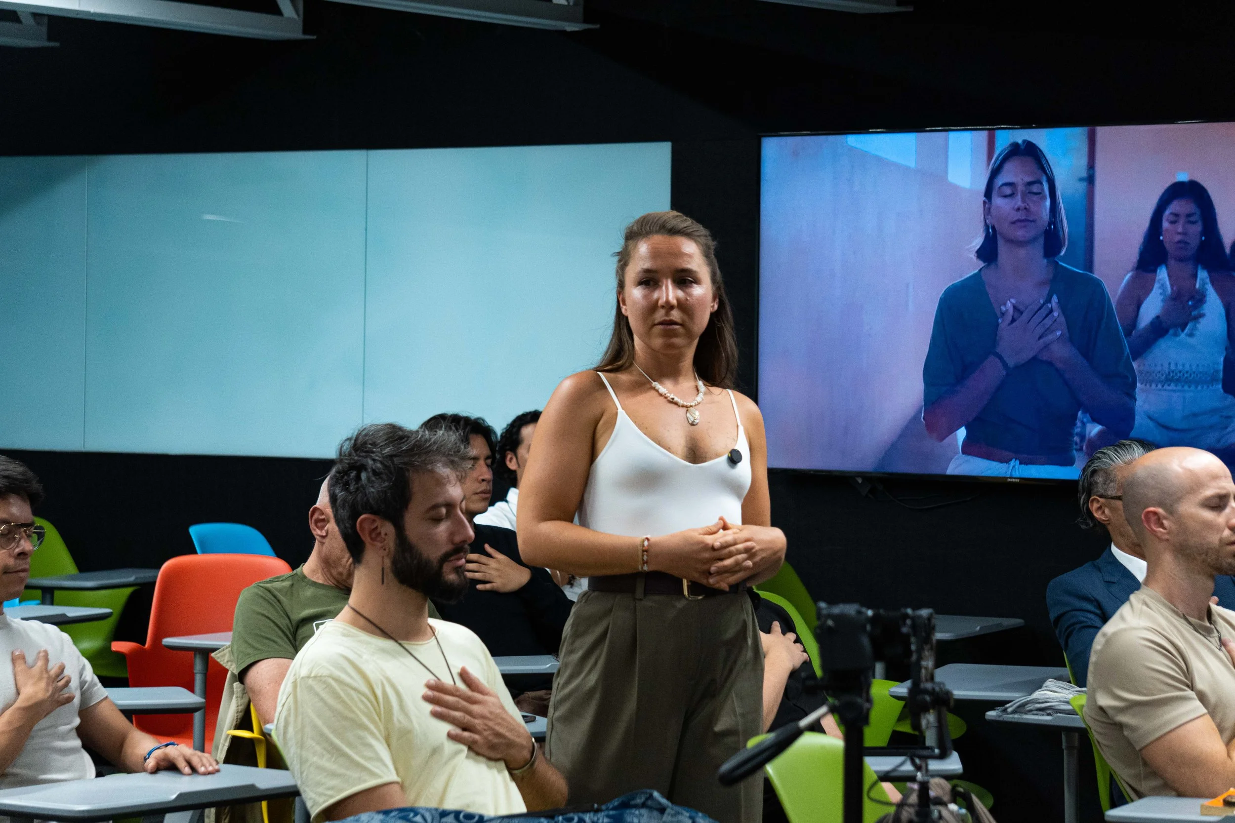 A woman in a white tank top and brown pants standing in a classroom, with her hand over her heart, surrounded by seated people with their hands over their hearts, and a large screen behind displaying her image.