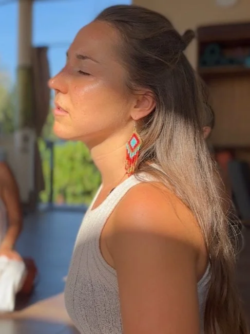 A woman with long, light brown hair tied back in a bun, wearing a sleeveless white top and colorful beaded earrings, sitting with her eyes closed in a sunlit room.