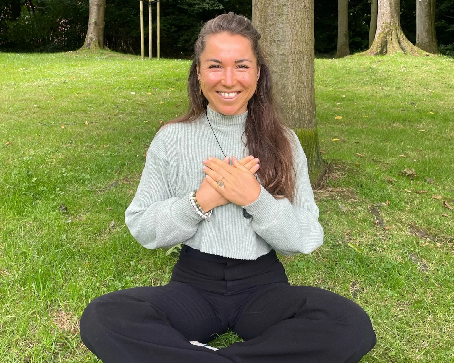 A woman sitting cross-legged on the grass in a park, smiling and holding her chest with both hands, wearing a gray turtleneck sweater and black pants, with trees in the background.