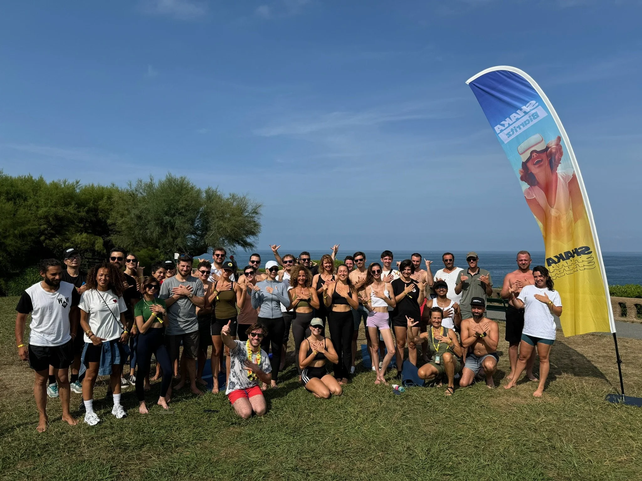 Group of people outdoors near the ocean, posing for a photo on a sunny day, with a blue sky, green trees, and a large yellow and blue flag with a woman wearing sunglasses.