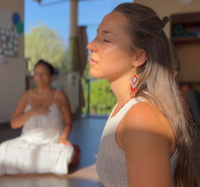 A woman in the foreground with her eyes closed, enjoying sunlight on her face, wearing a white sleeveless top and colorful earrings. Another woman in the background, blurred, wearing a white dress and sitting indoors by a window.
