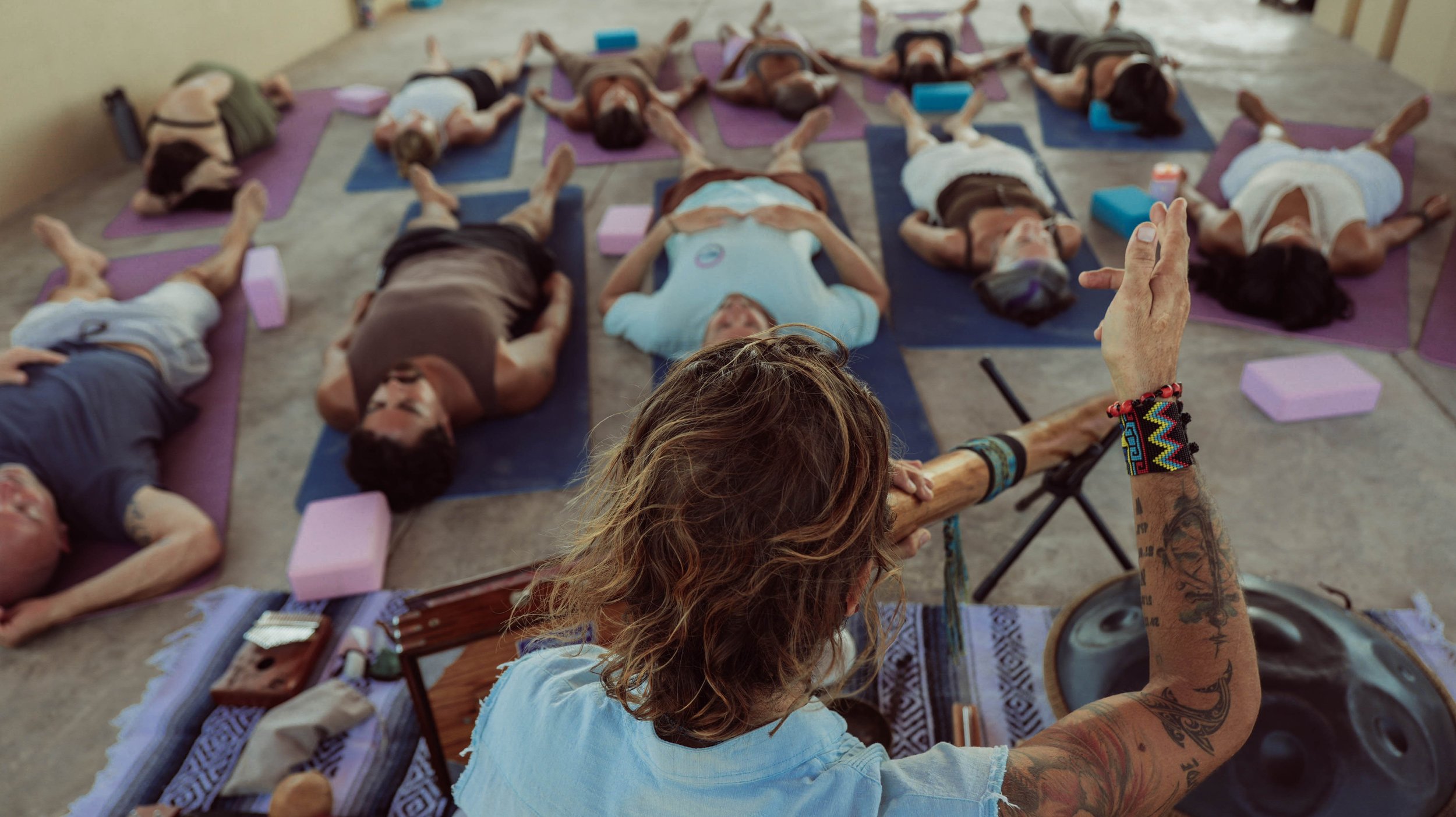Instructor leading a yoga or meditation class with multiple participants lying down on mats in a spacious indoor setting.