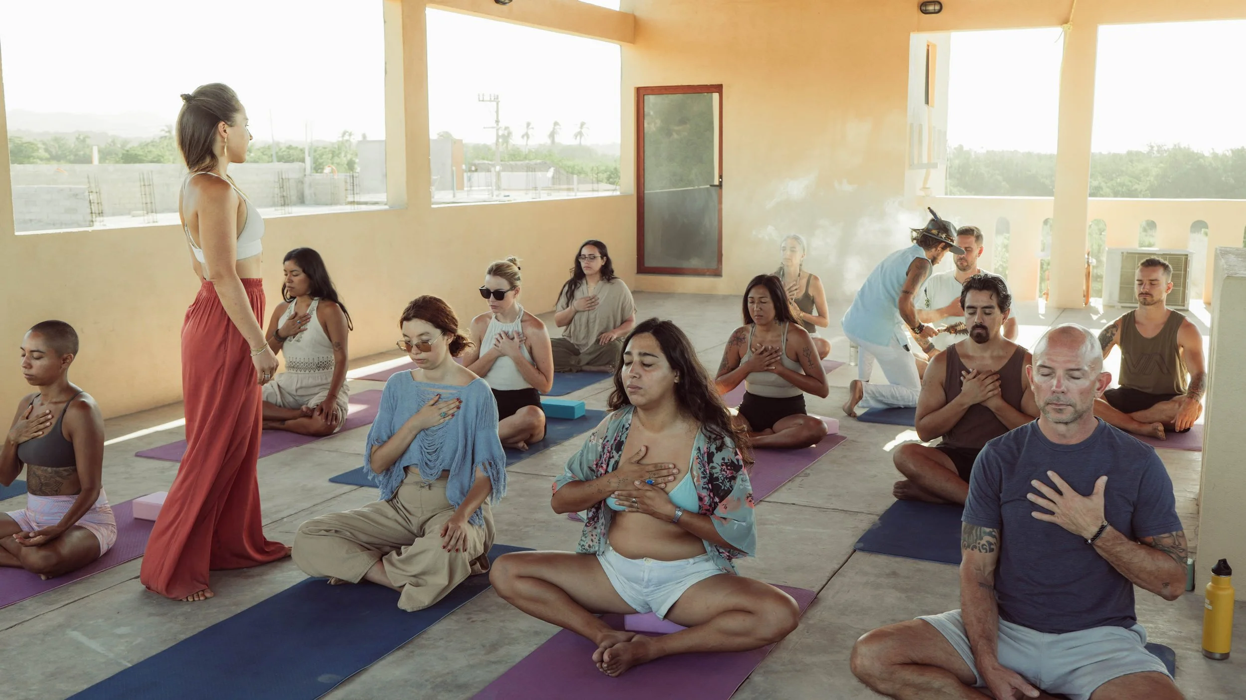 People practicing yoga on mats in a spacious, sunlit indoor space with large windows, some with their hands over their hearts and eyes closed, participating in a meditation or relaxation class.