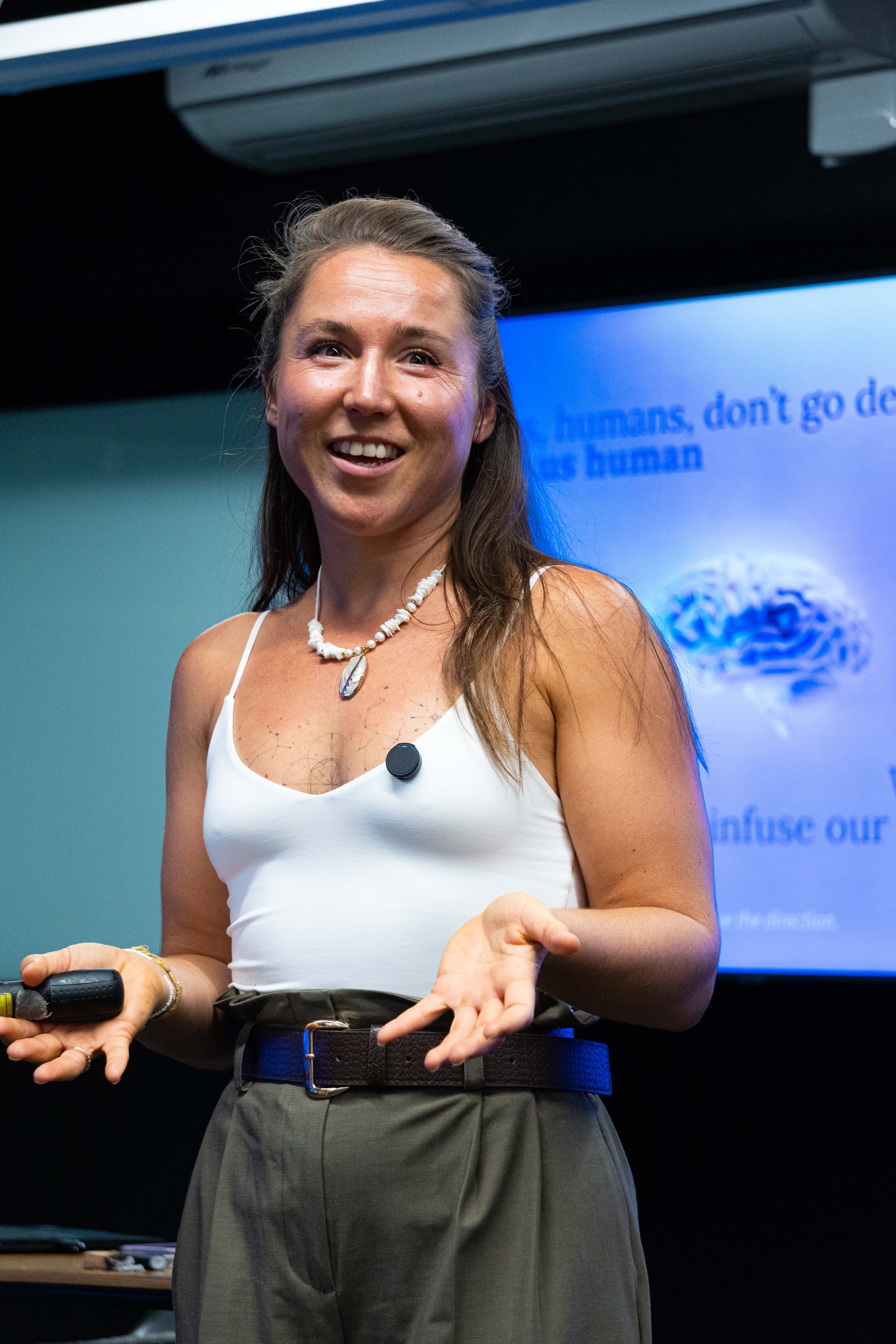 A woman with long brown hair, wearing a white tank top, khaki pants, a shell necklace, and a microphone clip on her top, giving a presentation in front of a screen displaying a slide about the human brain.