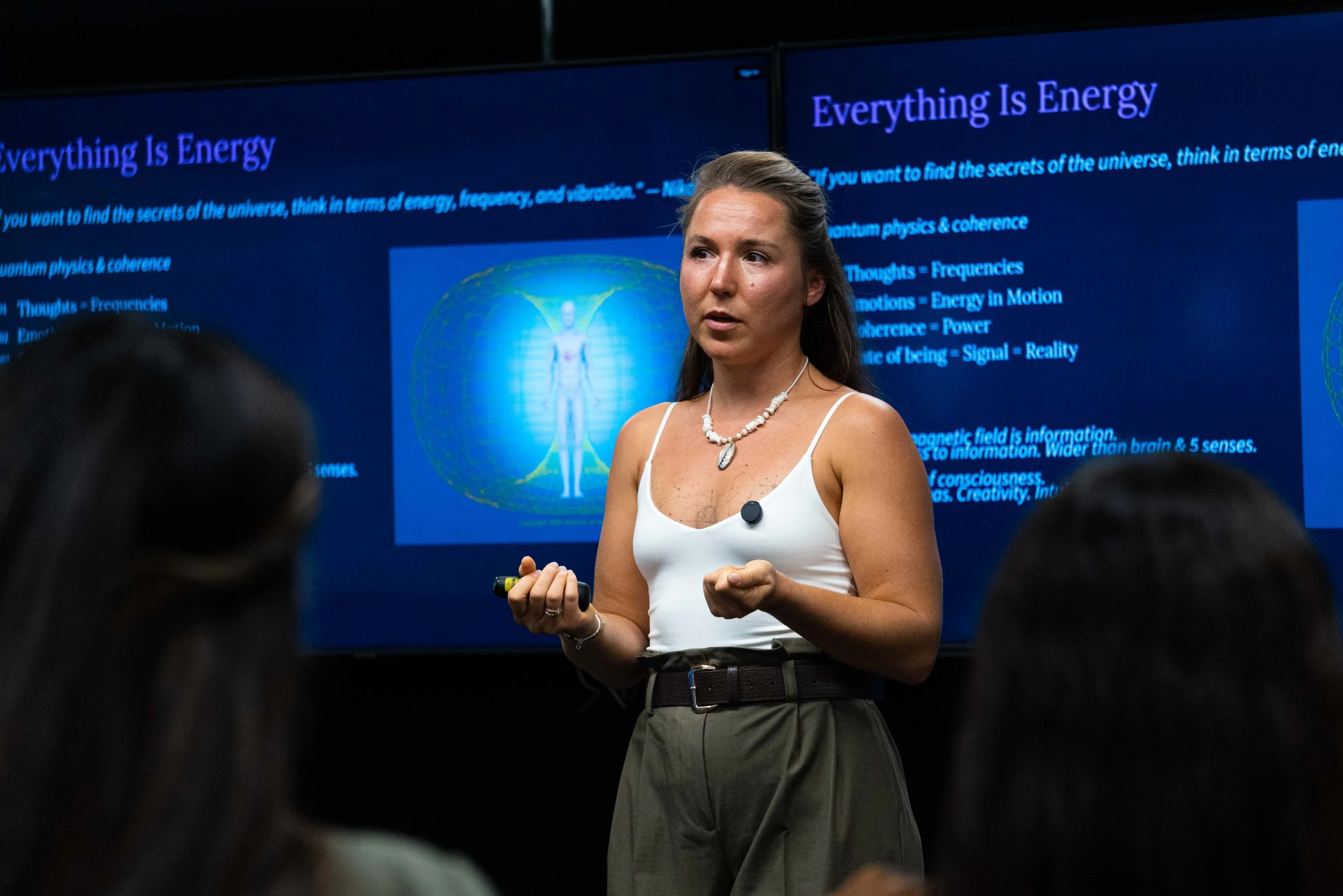 A woman giving a presentation in front of a large screen displaying a slide titled 'Everything Is Energy,' with an illustration of a human figure surrounded by a glowing energy field.