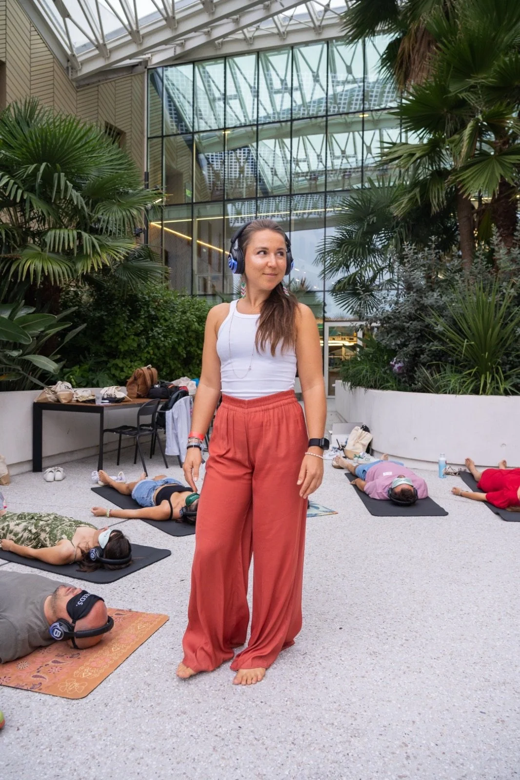 A woman sitting cross-legged on a mat outdoors, smiling, with lush green trees and hills in the background.