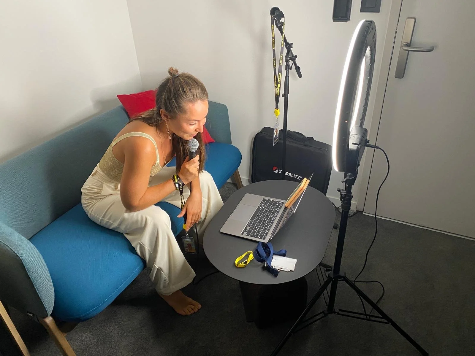 Woman sitting on a blue couch, holding a microphone, preparing to record or stream using a laptop in a small room with a bright ring light.