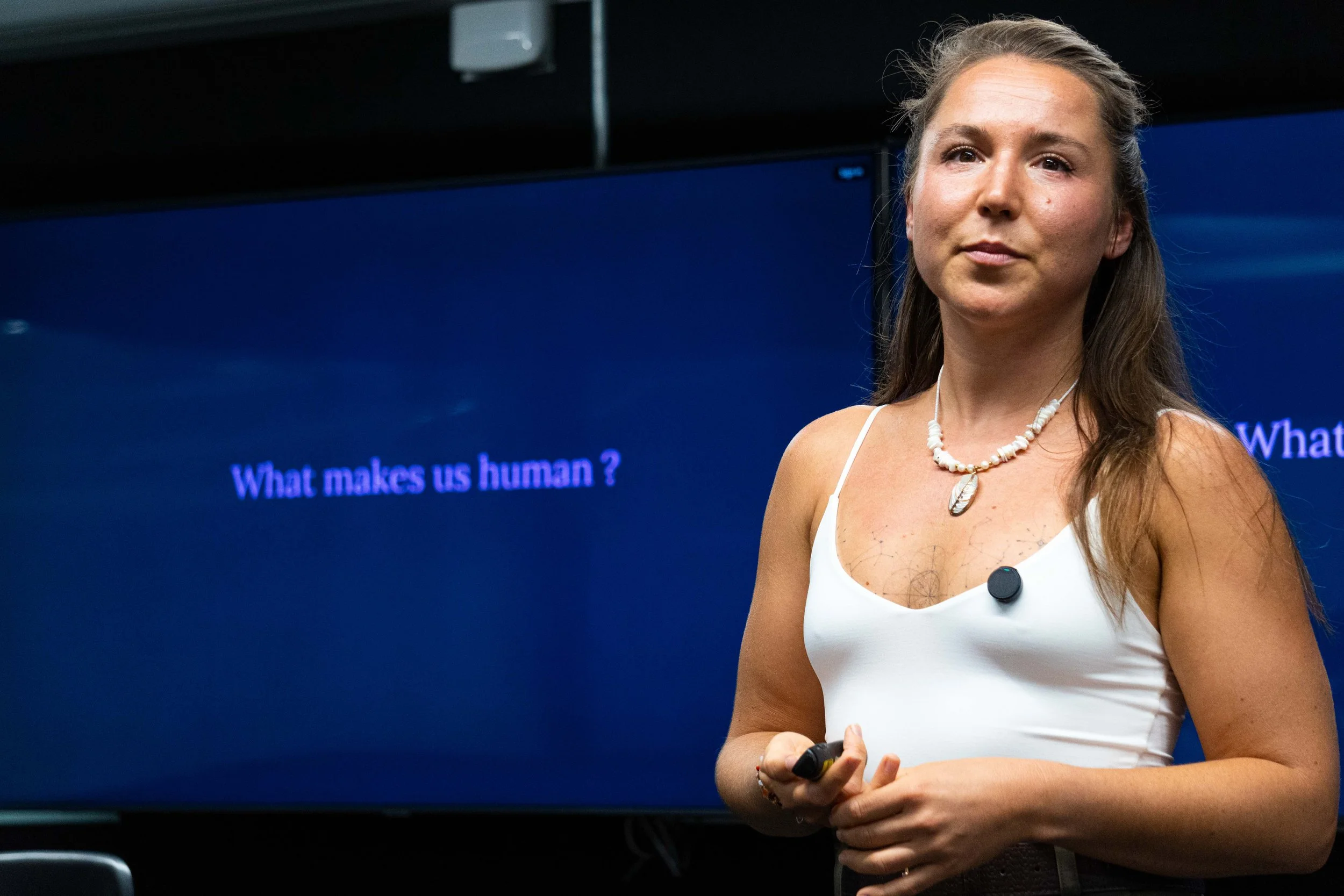 A woman wearing a white top and necklace standing in front of a blue screen with the text 'What makes us human?'
