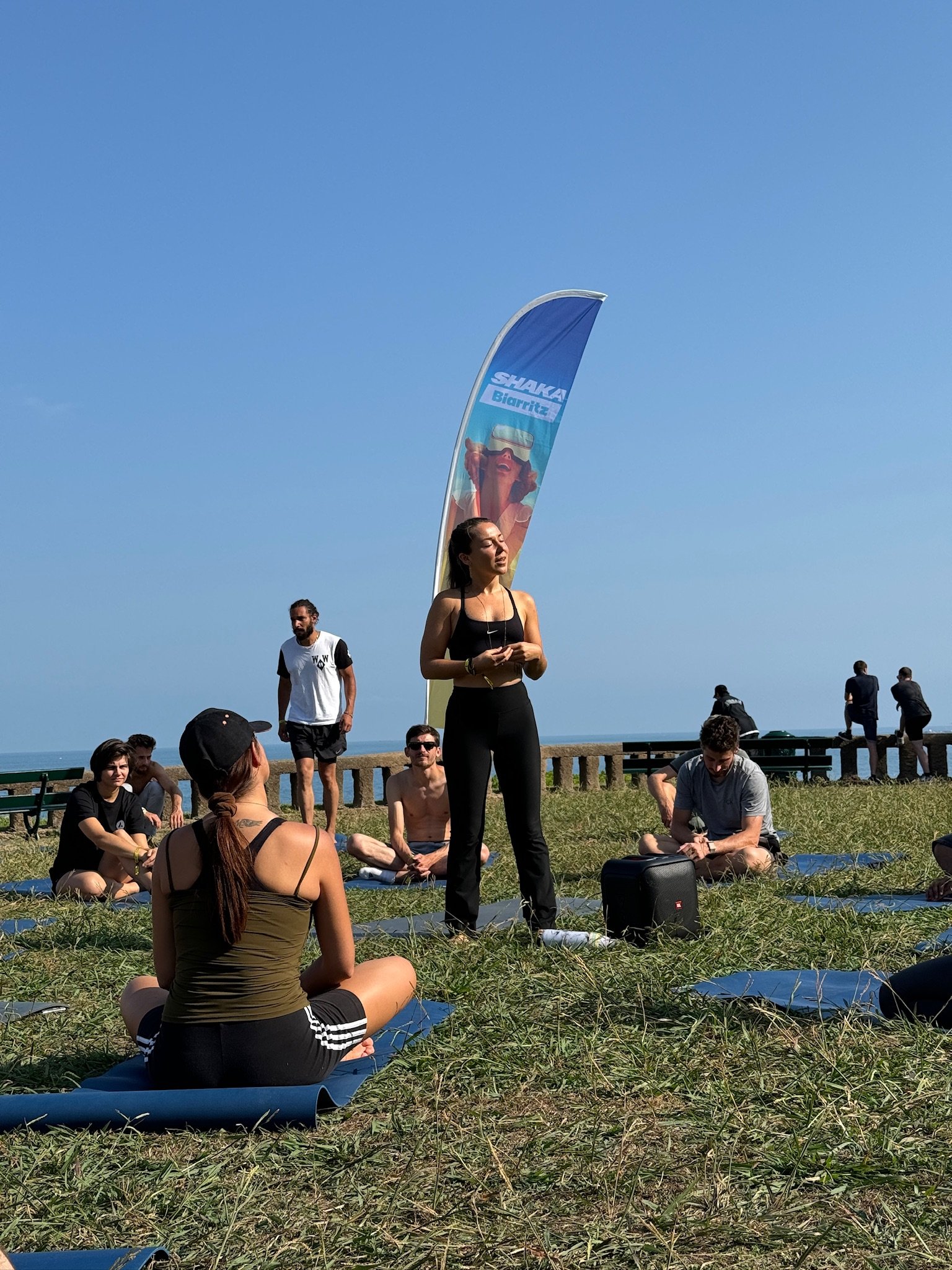 Group of people participating in outdoor yoga session on grass near the sea, with a woman leading the session, a banner, and a bright blue sky.