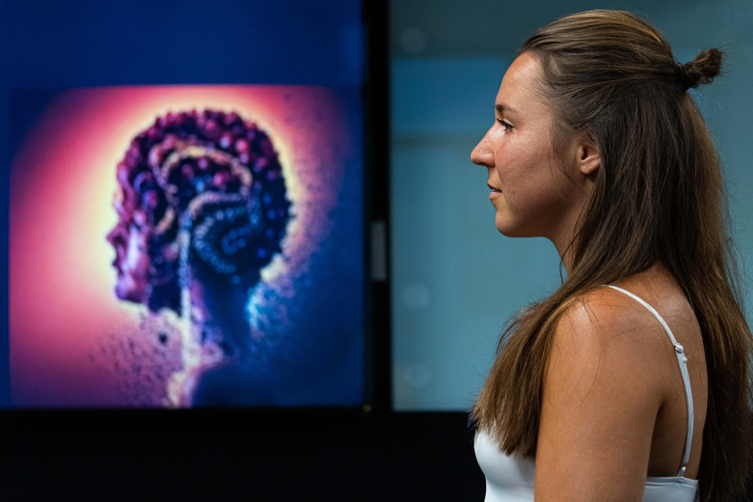 A woman with long brown hair, wearing a white tank top, standing in profile in front of a digital display showing a side profile of a person's head with a visible brain.