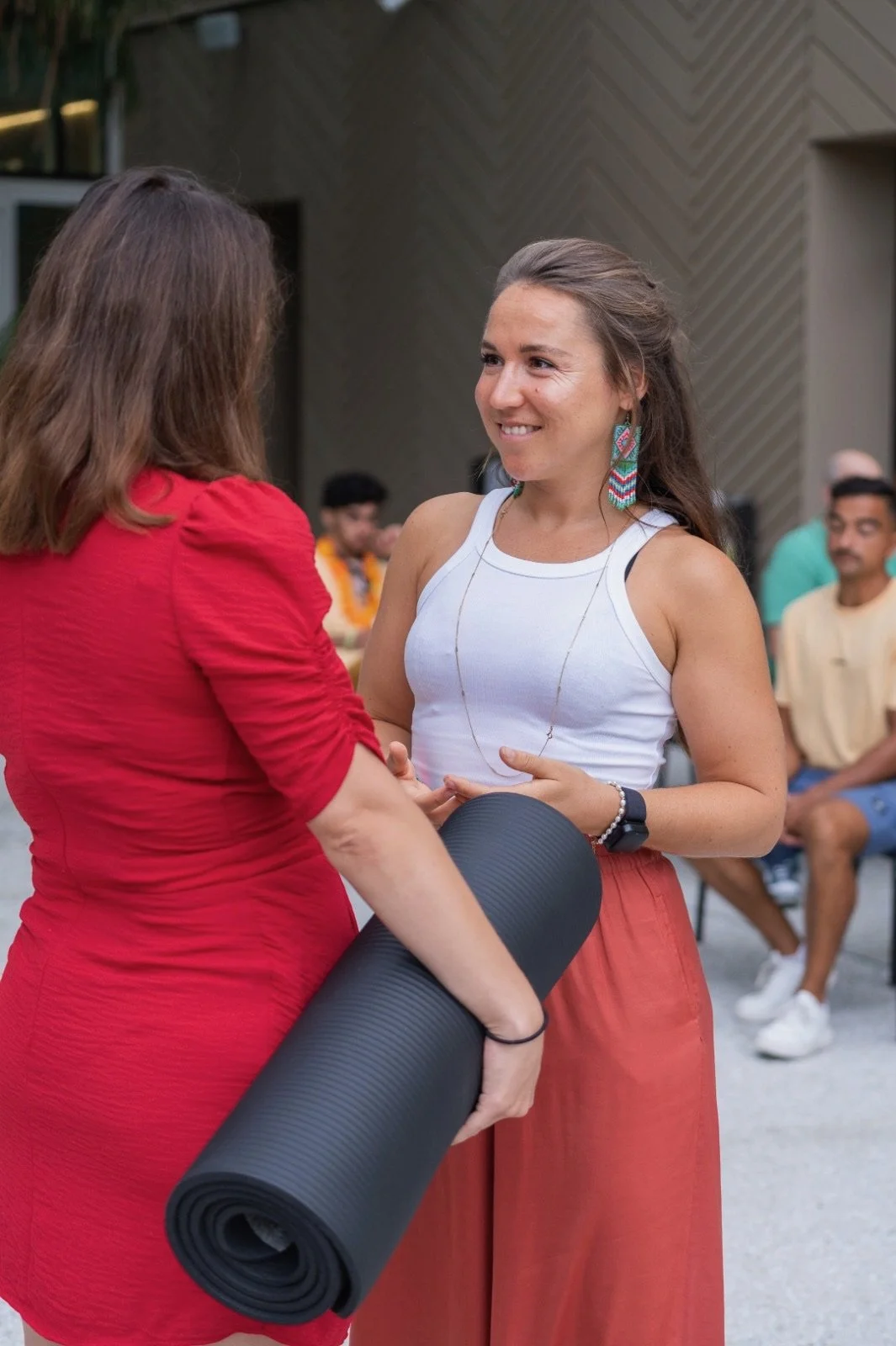 Two women are having a conversation outdoors, one holding a rolled yoga mat. The woman facing the camera has long brown hair, is smiling, and is wearing a white sleeveless top, earrings, and a watch. The woman with her back to the camera has shoulder-length brown hair and is wearing a red dress. In the background, there are a few people sitting and standing, with a modern building wall visible.